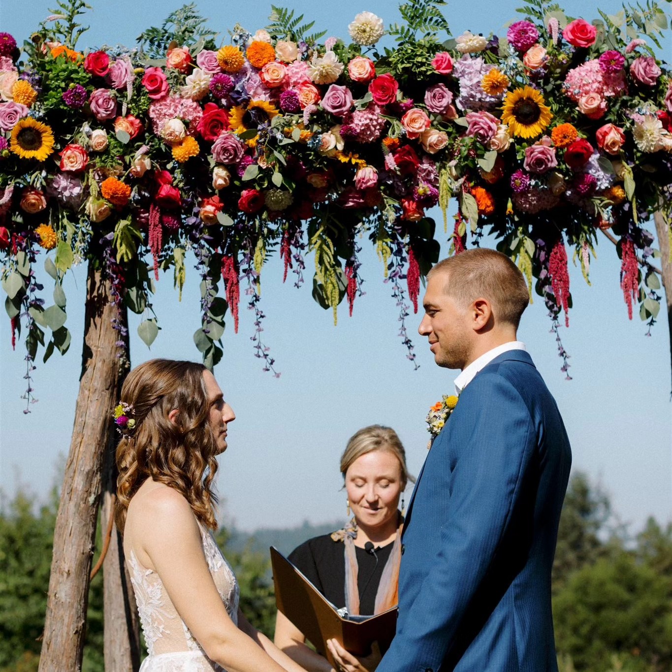 A wedding ceremony with a couple facing each other under a floral arch, with an officiant holding a book, outdoors.