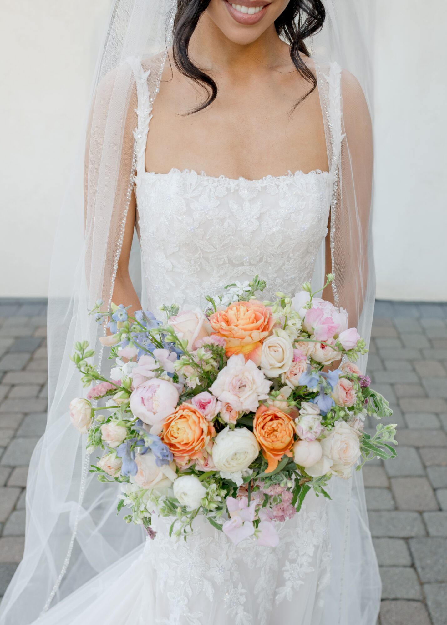 Bride in wedding dress holding a large bouquet of pink, orange, and white flowers, smiling, outdoors.