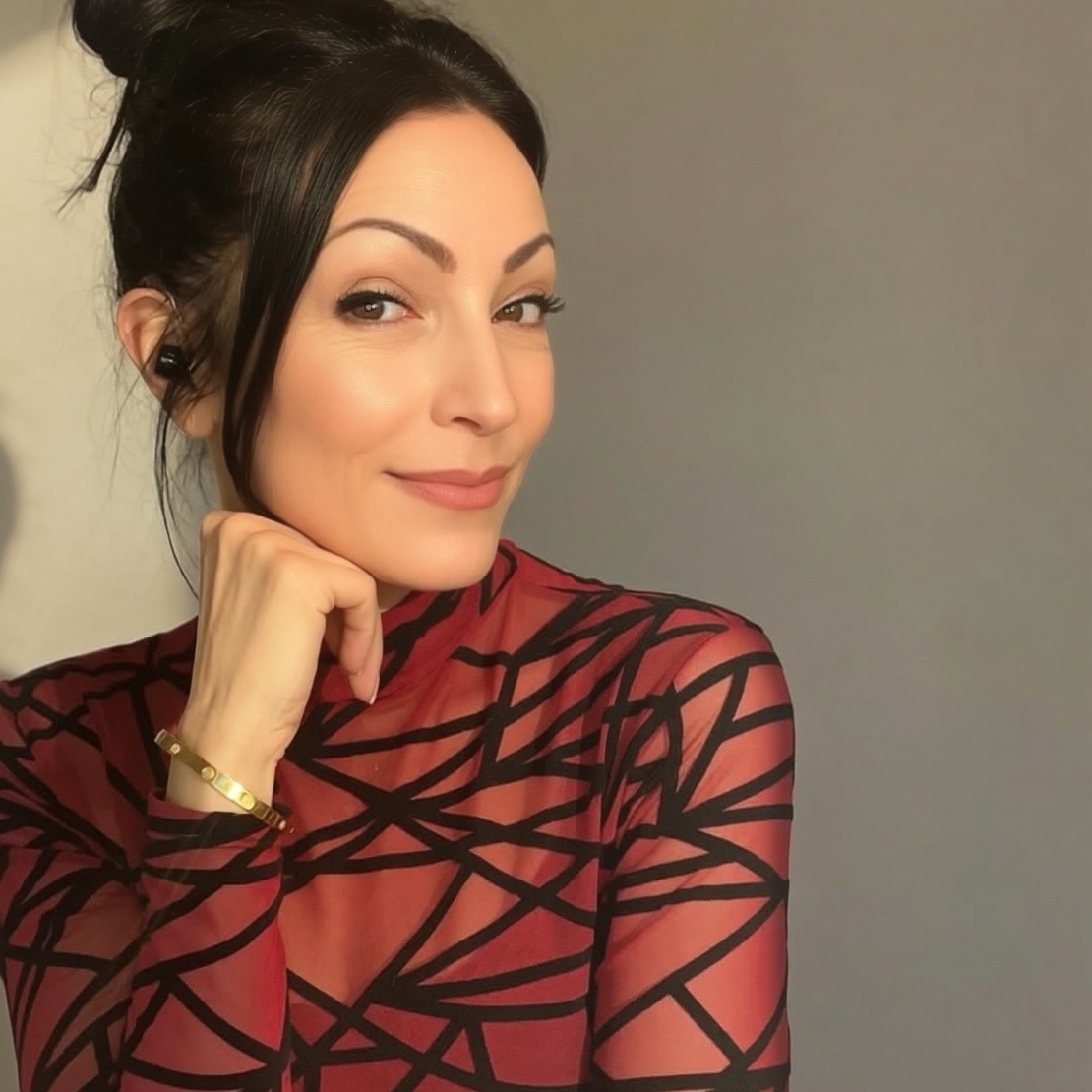 Woman with dark hair in a bun, wearing a patterned top, smiling, resting her chin on her hand, against a plain background.
