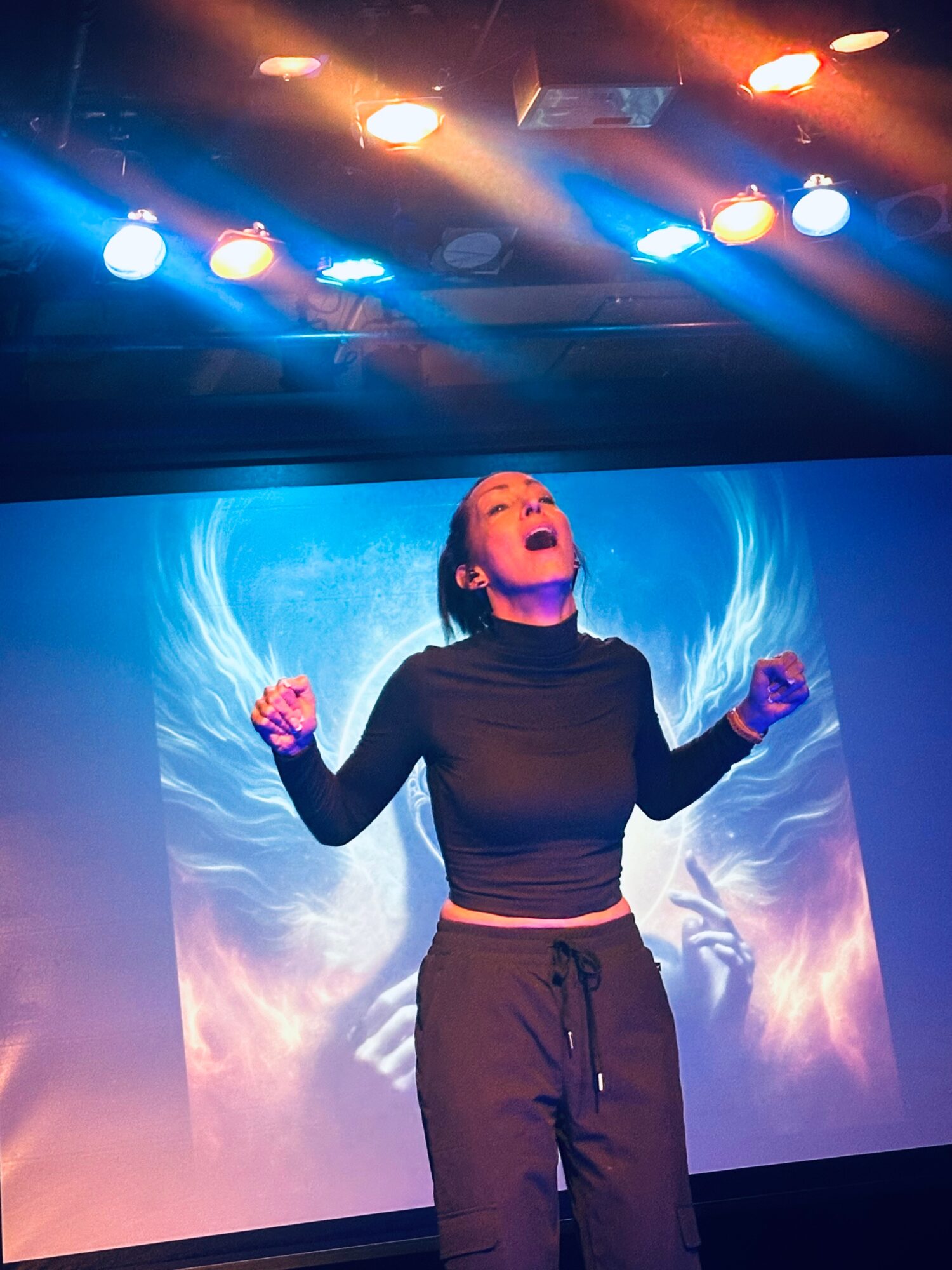 A woman stands on stage with arms raised, singing with eyes closed, illuminated by colorful stage lights and a blue background.