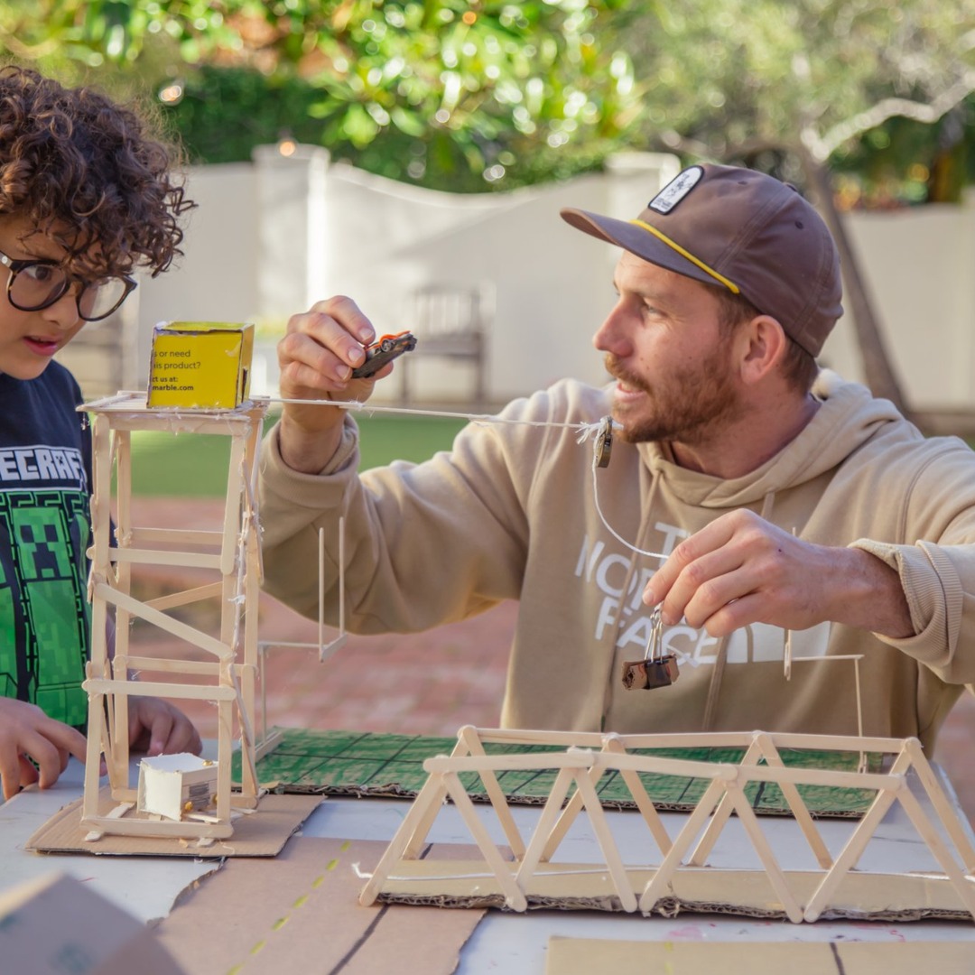 Two people building a wooden structure outdoors, with trees and a fence in the background.