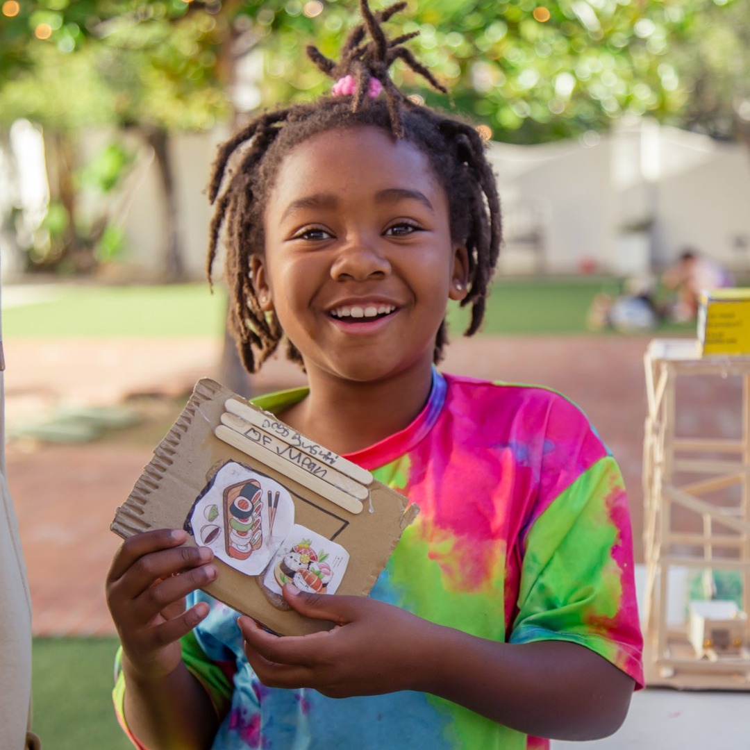 Smiling girl with dreadlocks holding a decorated cardboard box outdoors, wearing a colorful tie-dye shirt.