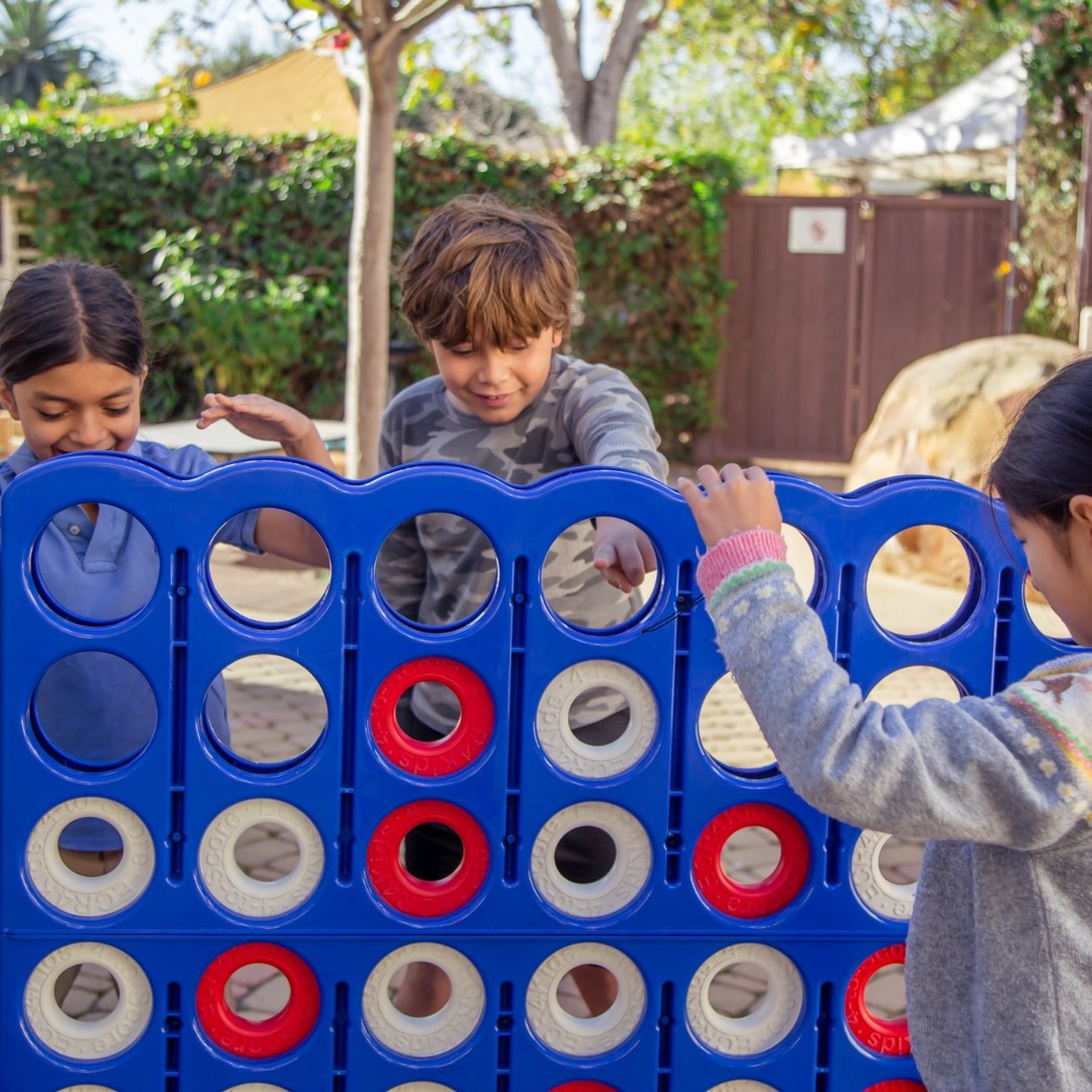 Three children playing Connect Four outdoors, with trees and a fence in the background.