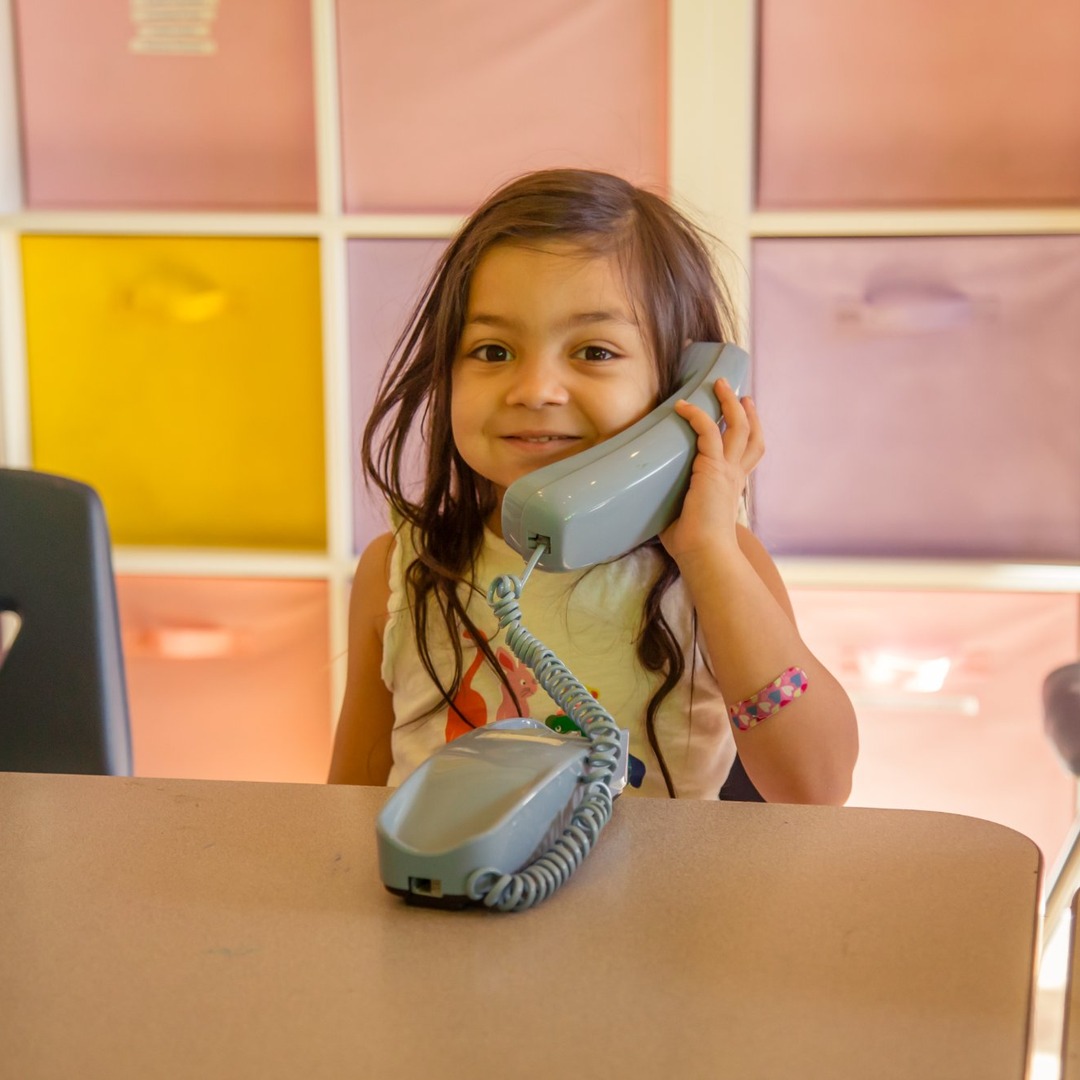 Young girl with long hair talking on a gray landline phone, colorful background with storage bins, smiling.
