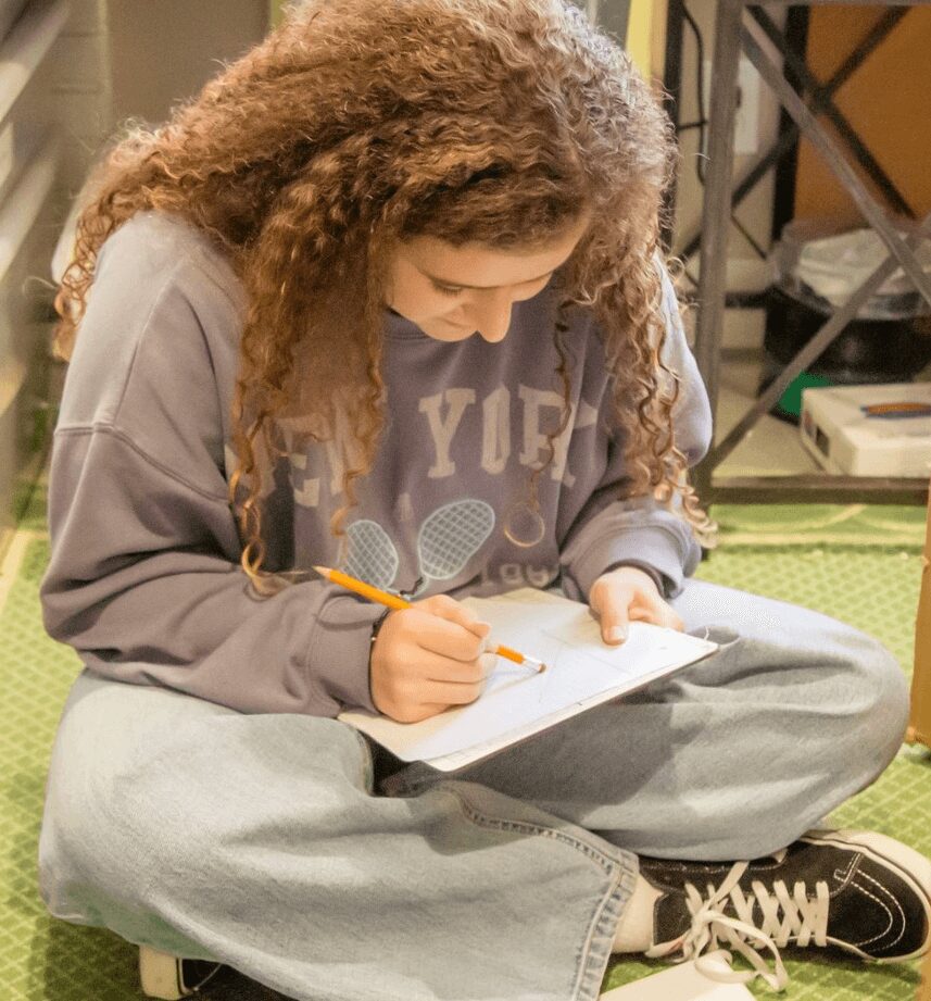 Person with curly hair sitting cross-legged, writing in a notebook with a pencil, in a room with shelves and books.