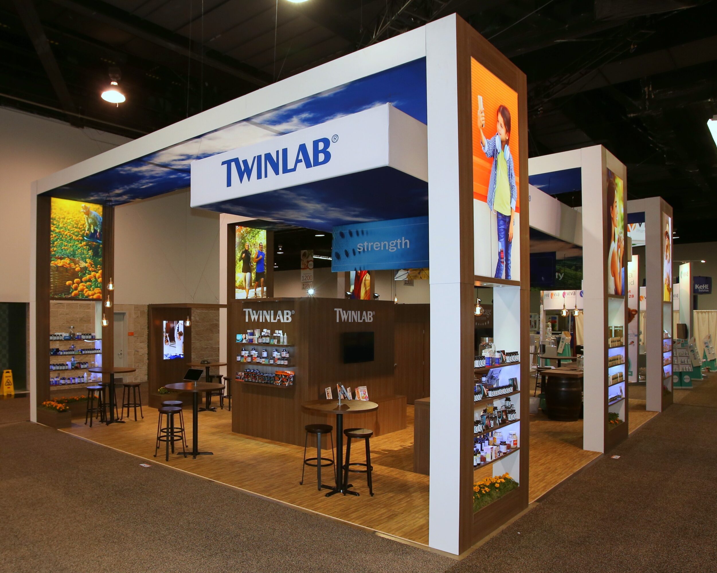 Trade show booth with Twinlab branding, display cases, and seating area, inside an exhibition hall with high ceilings.