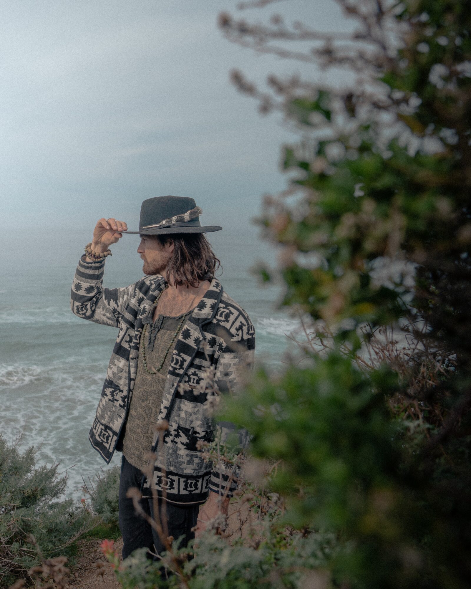 Person wearing a hat and patterned sweater standing near the ocean with cloudy sky, partially obscured by greenery.