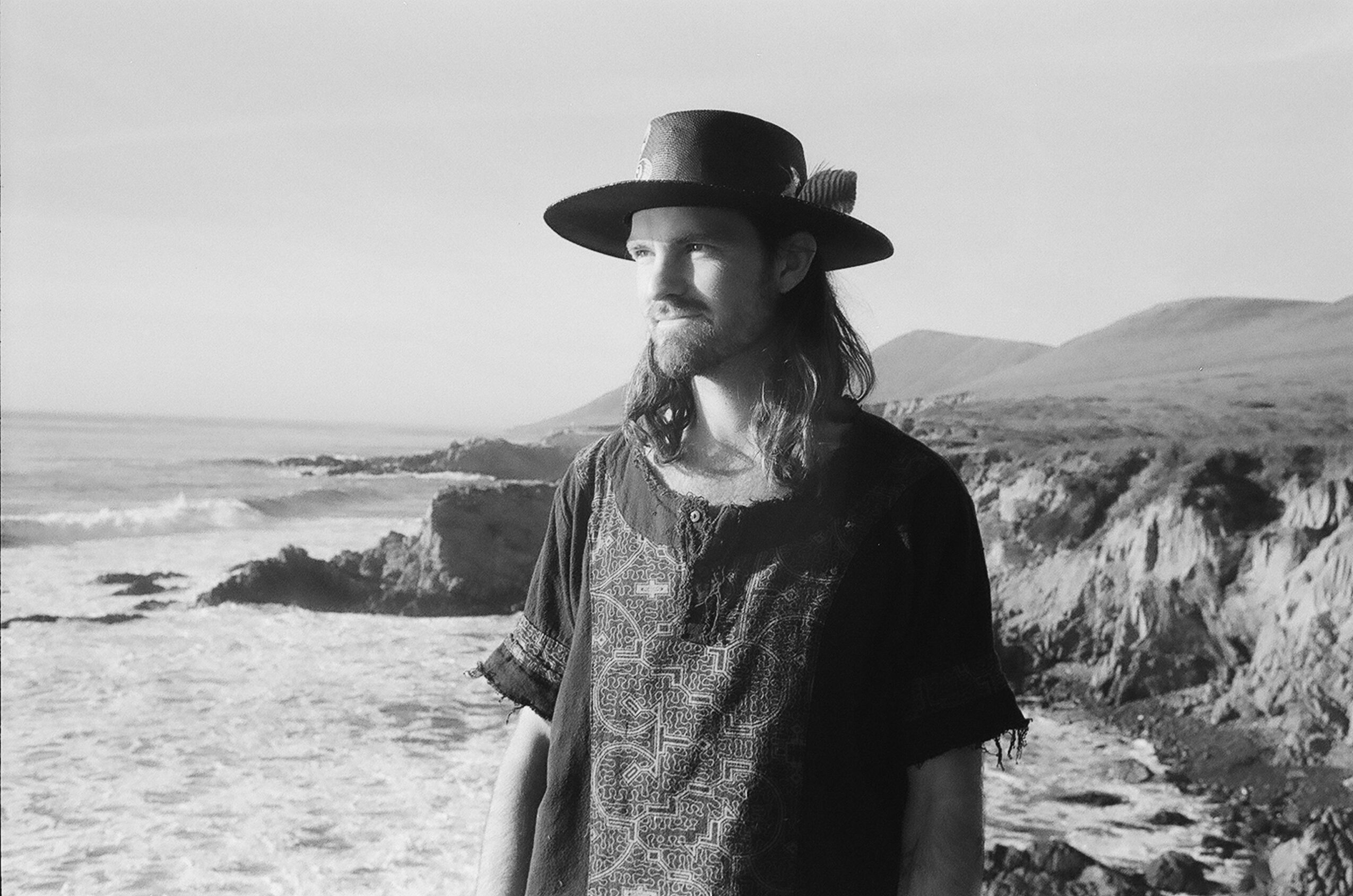Man with long hair and beard wearing a wide-brimmed hat and patterned shirt stands on a rocky beach with cliffs in background.