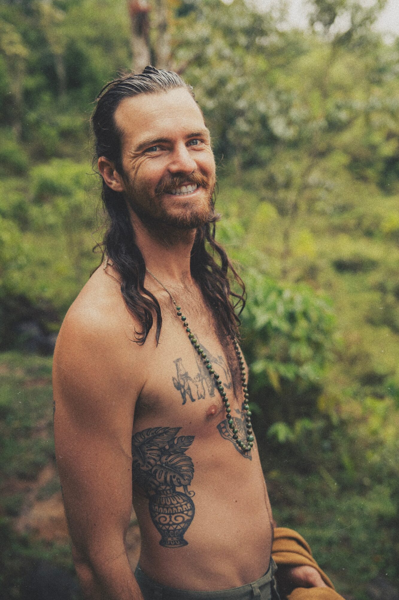 Man with long hair and beard smiling outdoors, shirtless, with tattoos and beaded necklace, surrounded by greenery.