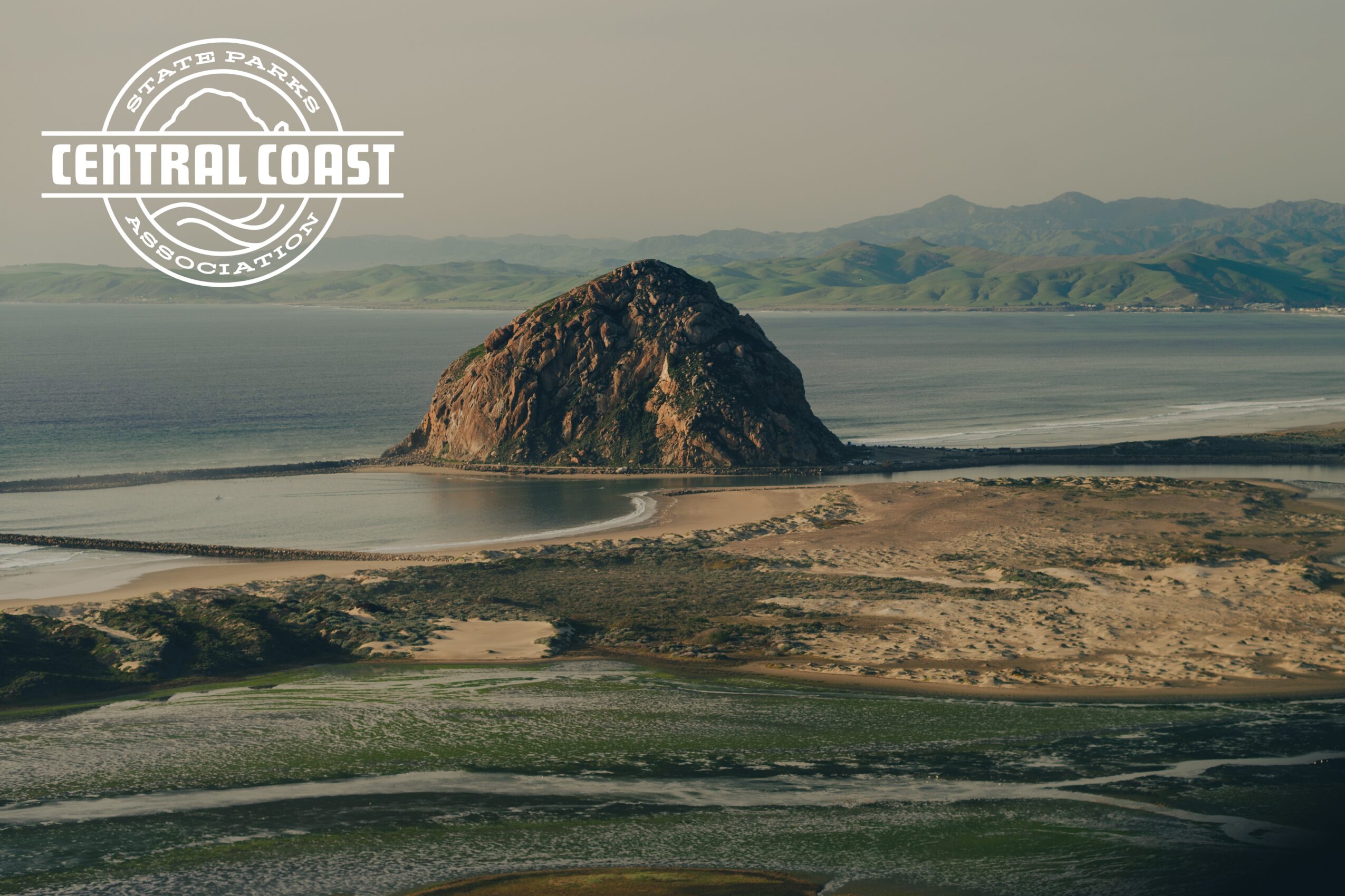 A large rock formation in a body of water with land and mountains in the background, overcast sky, Central Coast logo.
