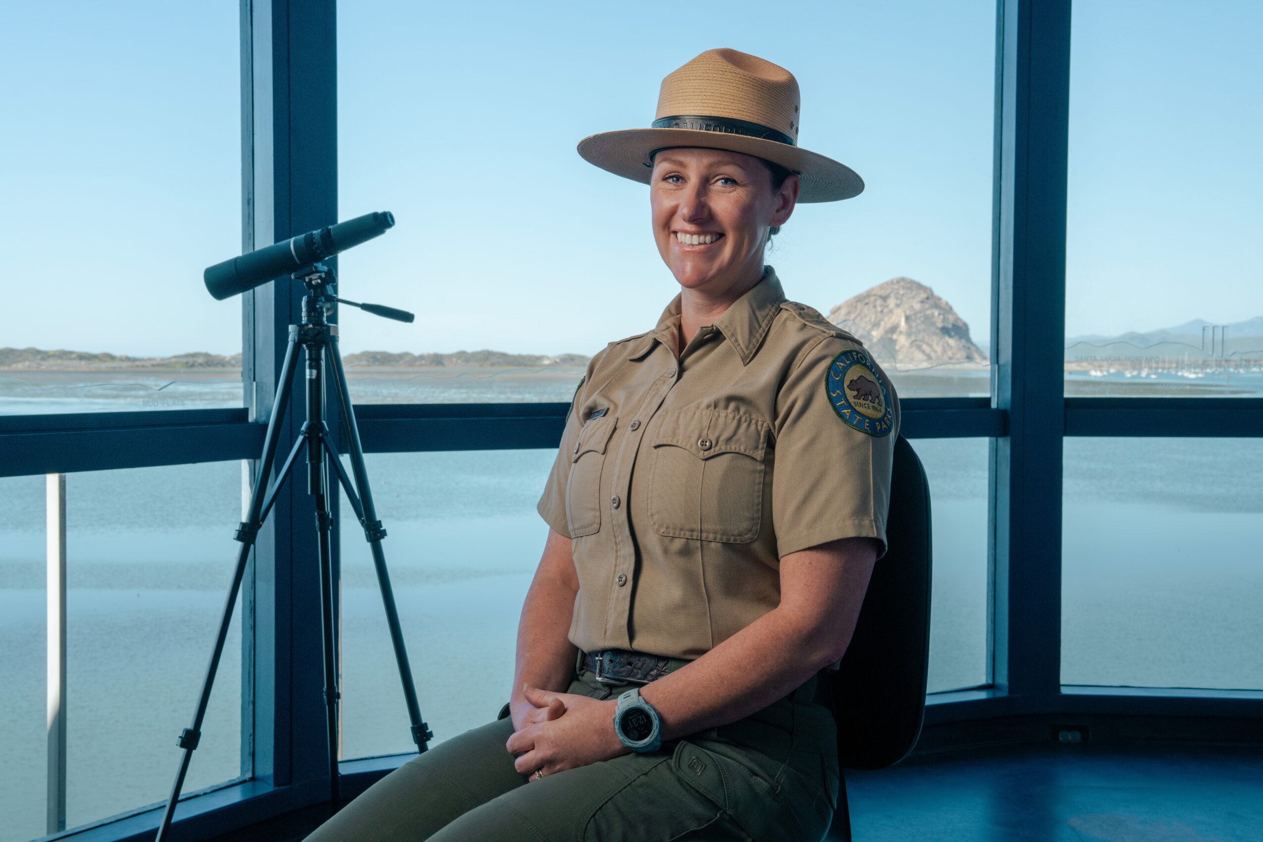 Female park ranger sitting indoors with a view of water and mountains, smiling, wearing a uniform and hat.