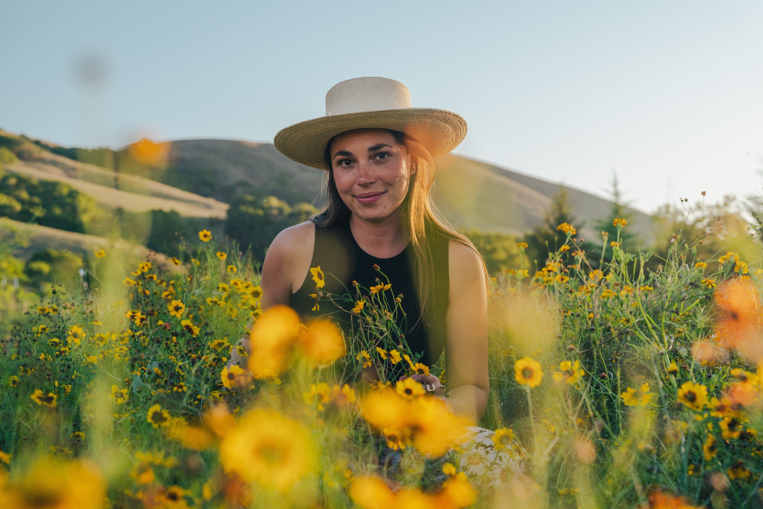 Young woman with a wide-brimmed hat smiling in a field of yellow flowers, hills in the background, sunny day.