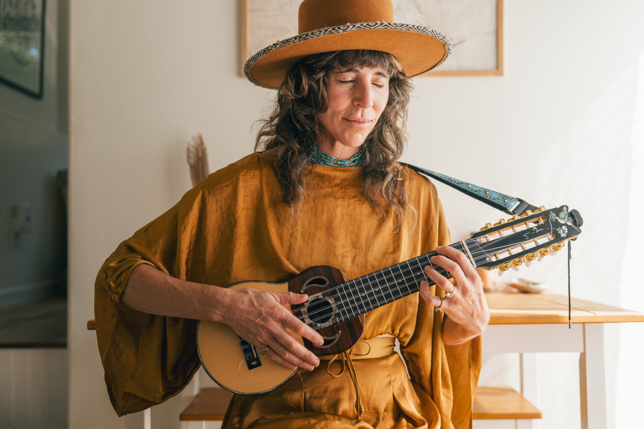 Woman wearing a large hat and brown outfit playing a small guitar indoors, with a light-colored wall and window in the background.