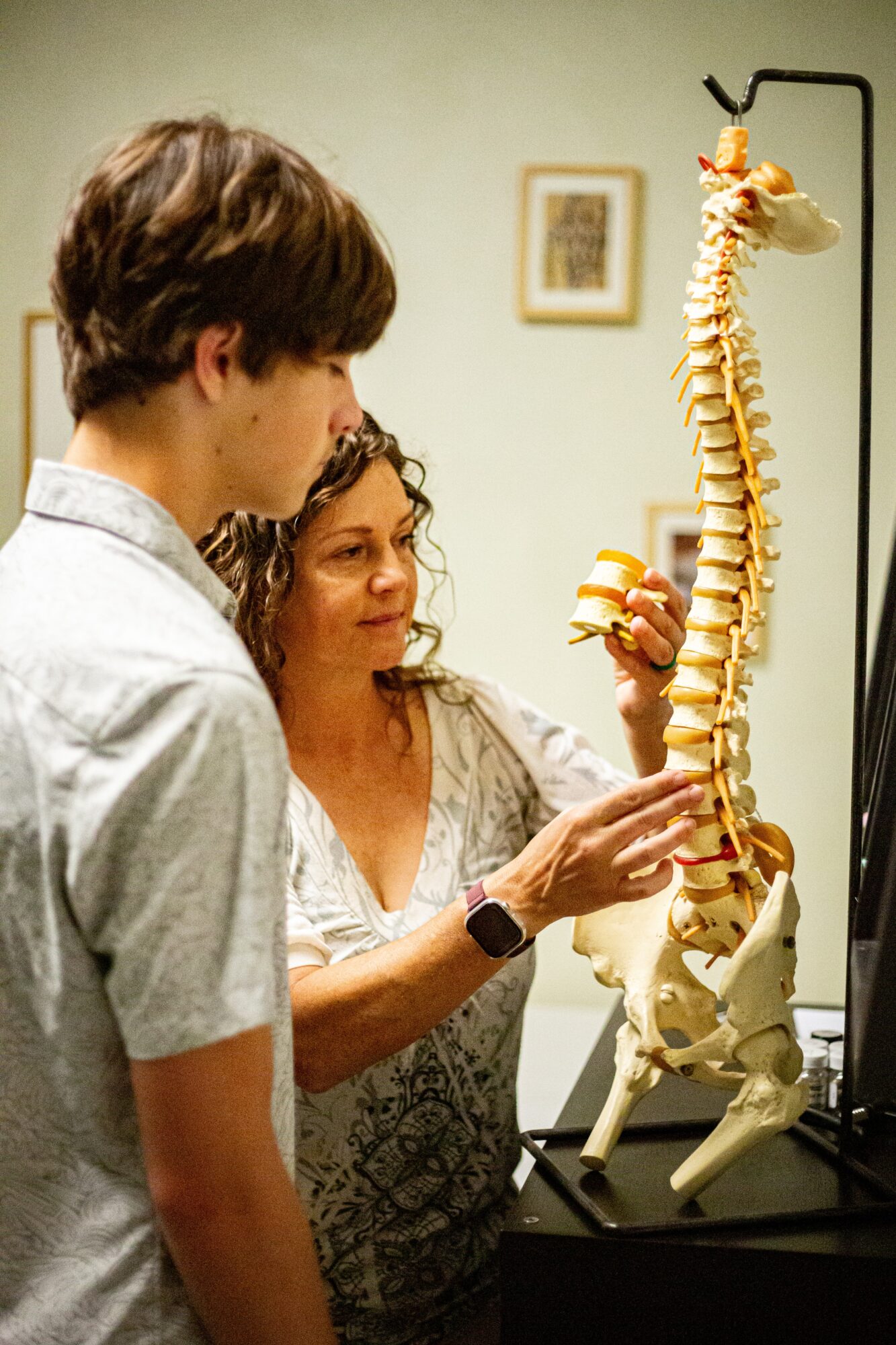 Woman and young man examining a human spine model in a medical setting, with framed pictures on the wall behind them.