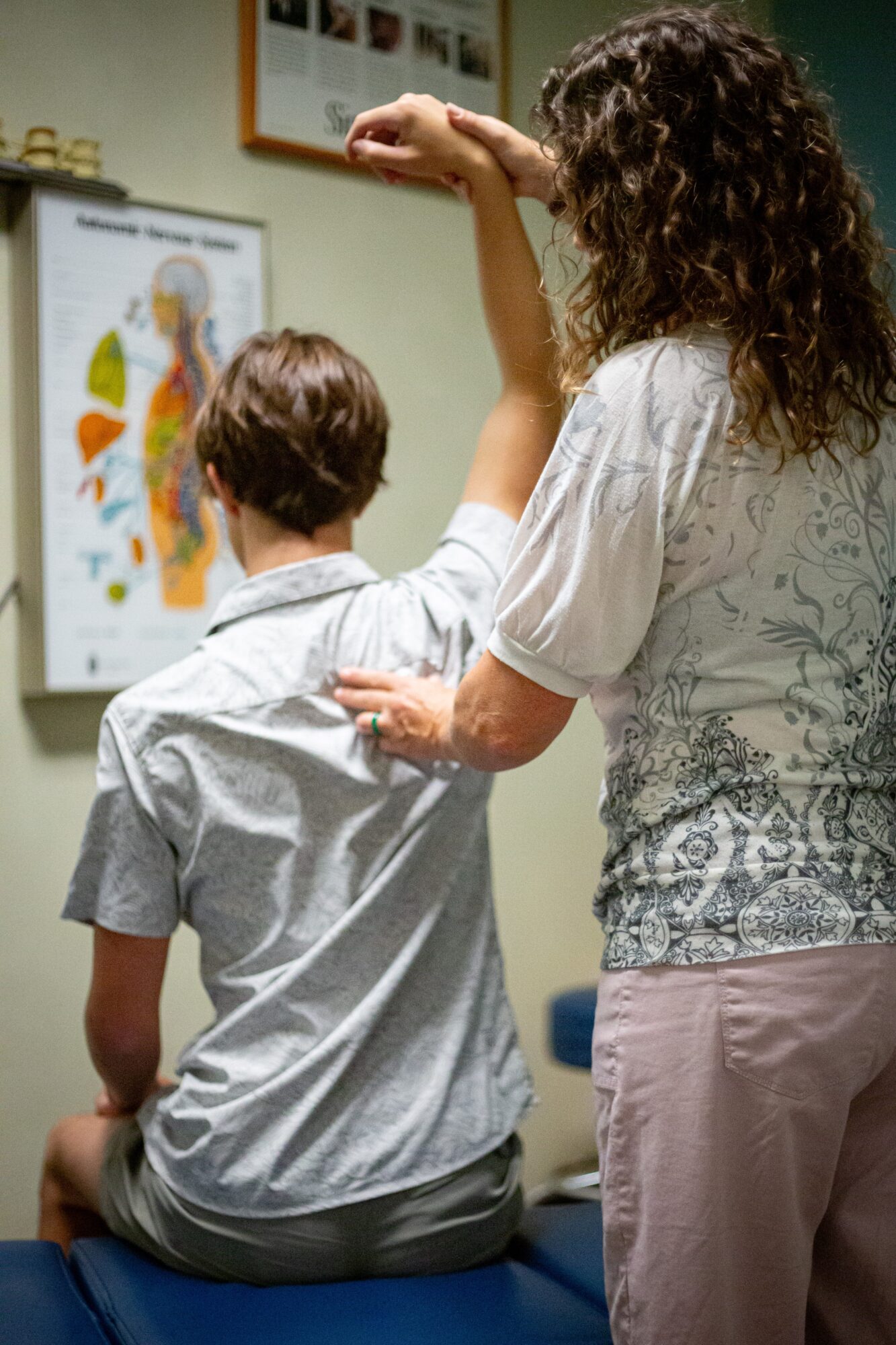 Two people, one sitting and one standing, looking at an anatomical poster on a wall.