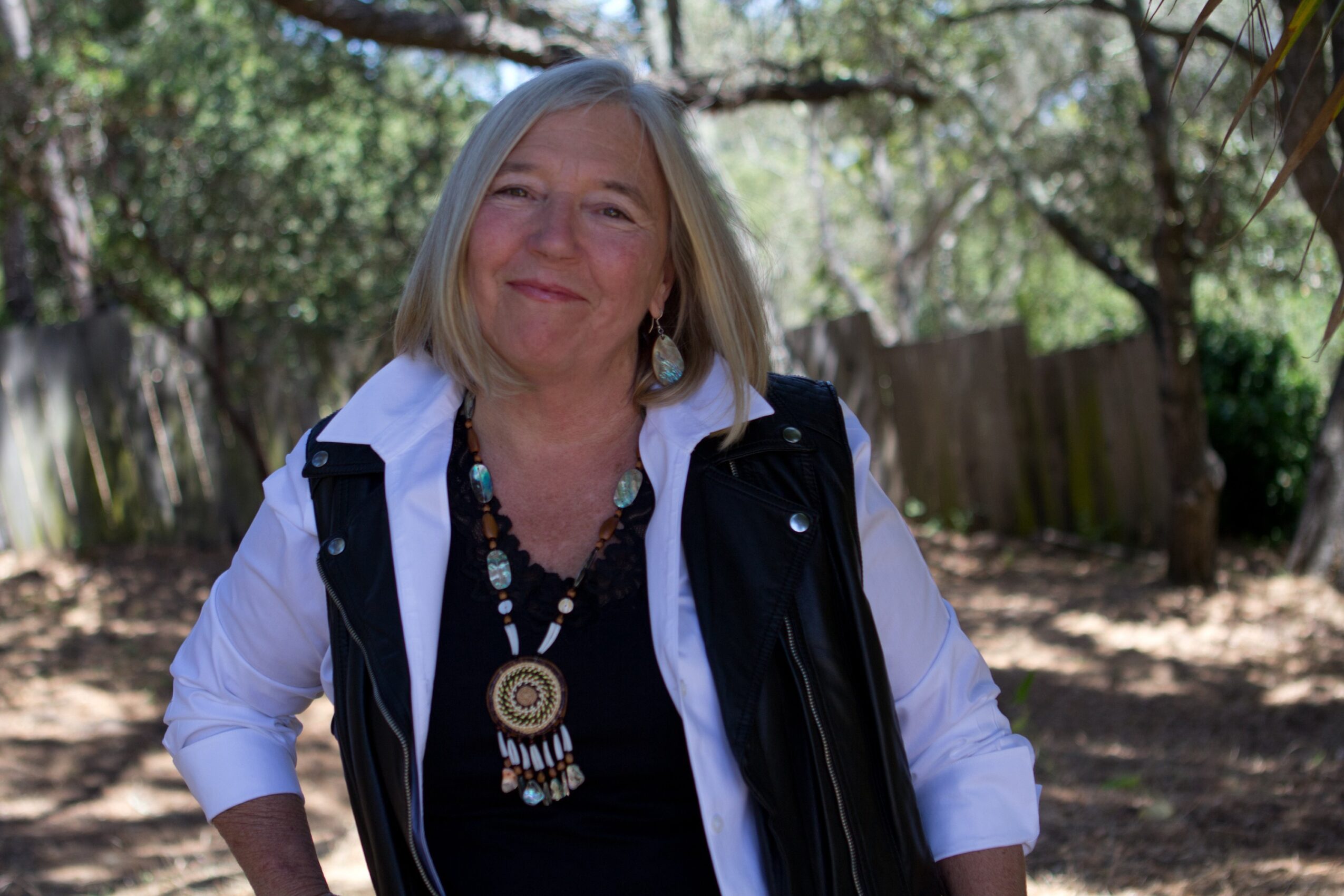A woman with shoulder-length gray hair smiling outdoors, wearing a white shirt, black vest, and colorful jewelry.