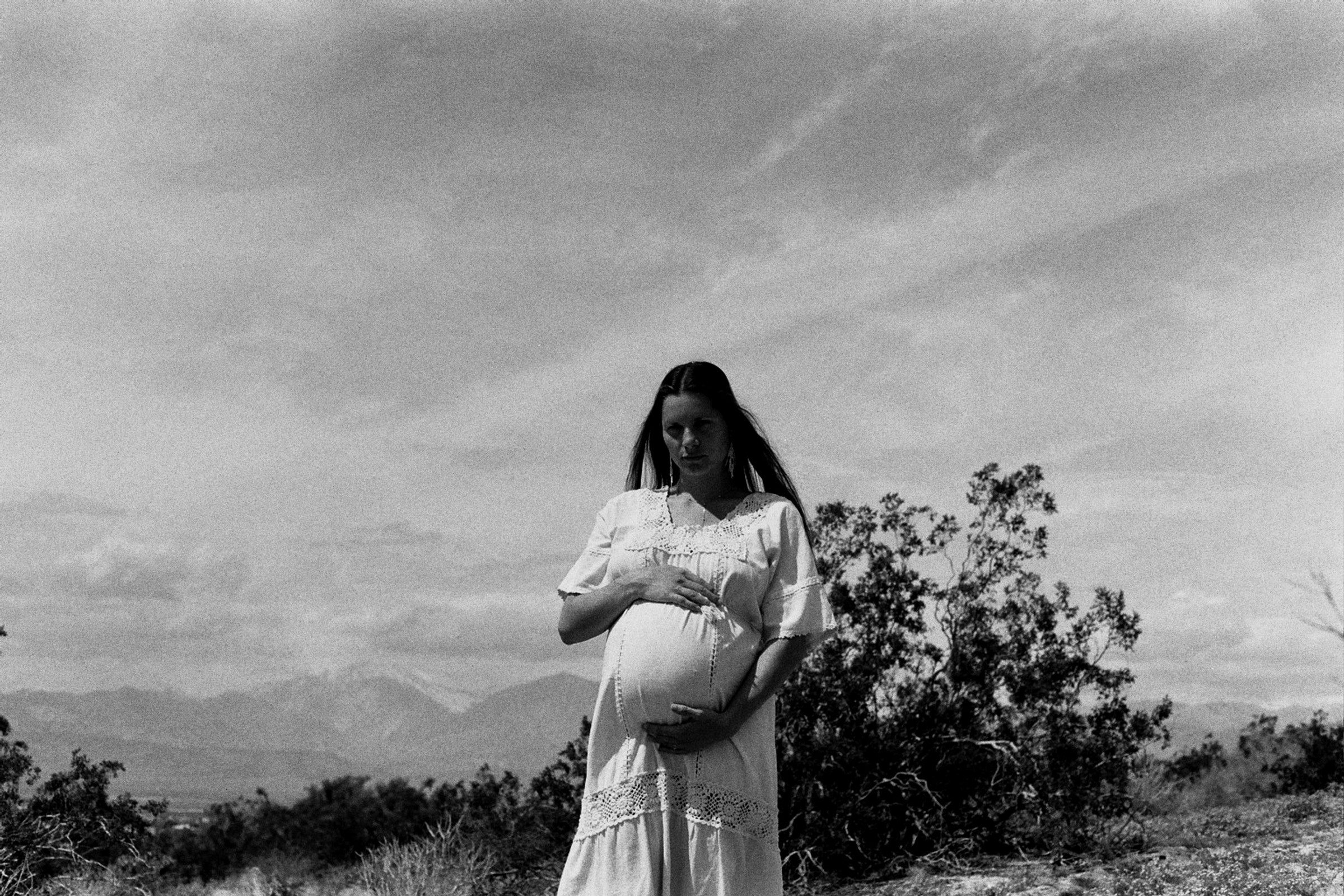 Woman in a long dress holding a pregnant belly outdoors, with trees and sky in background.