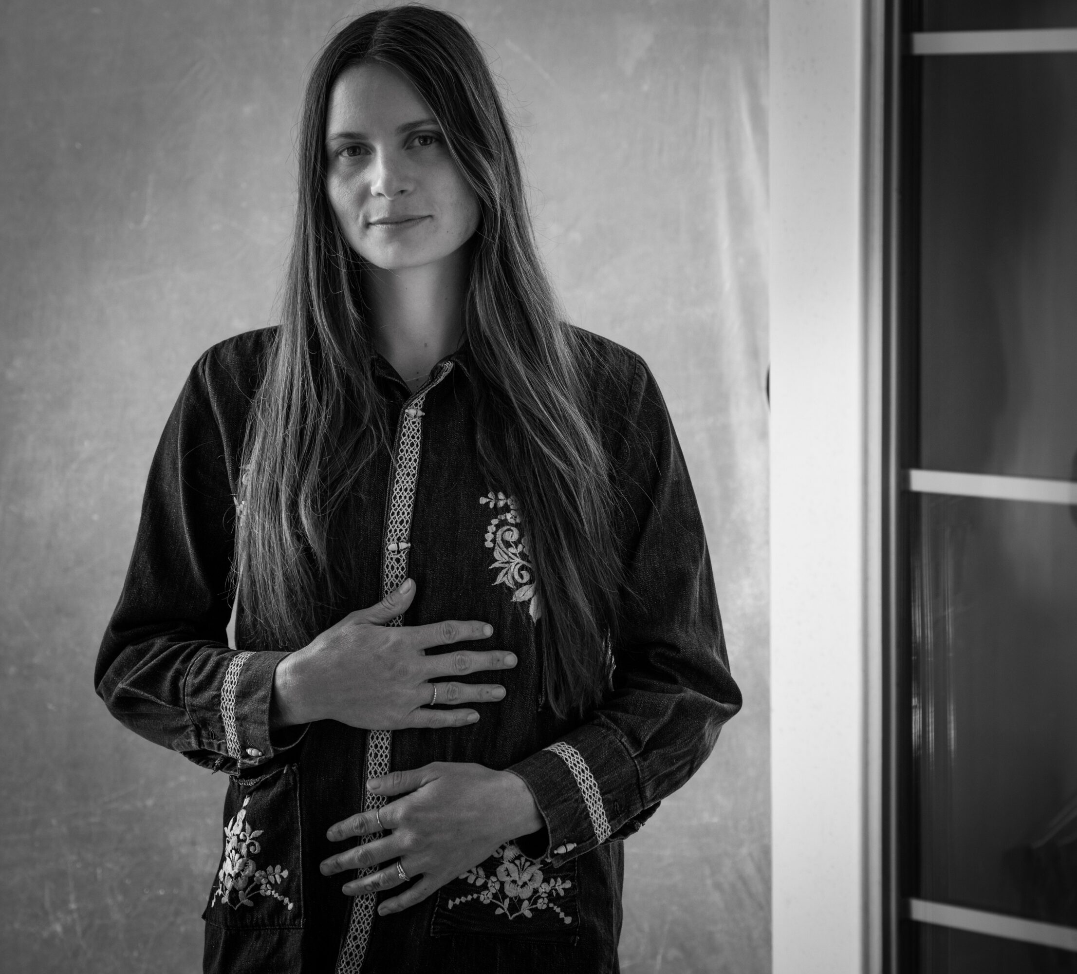 Young woman with long hair standing indoors, wearing a dark jacket, hand on chest, looking at camera.