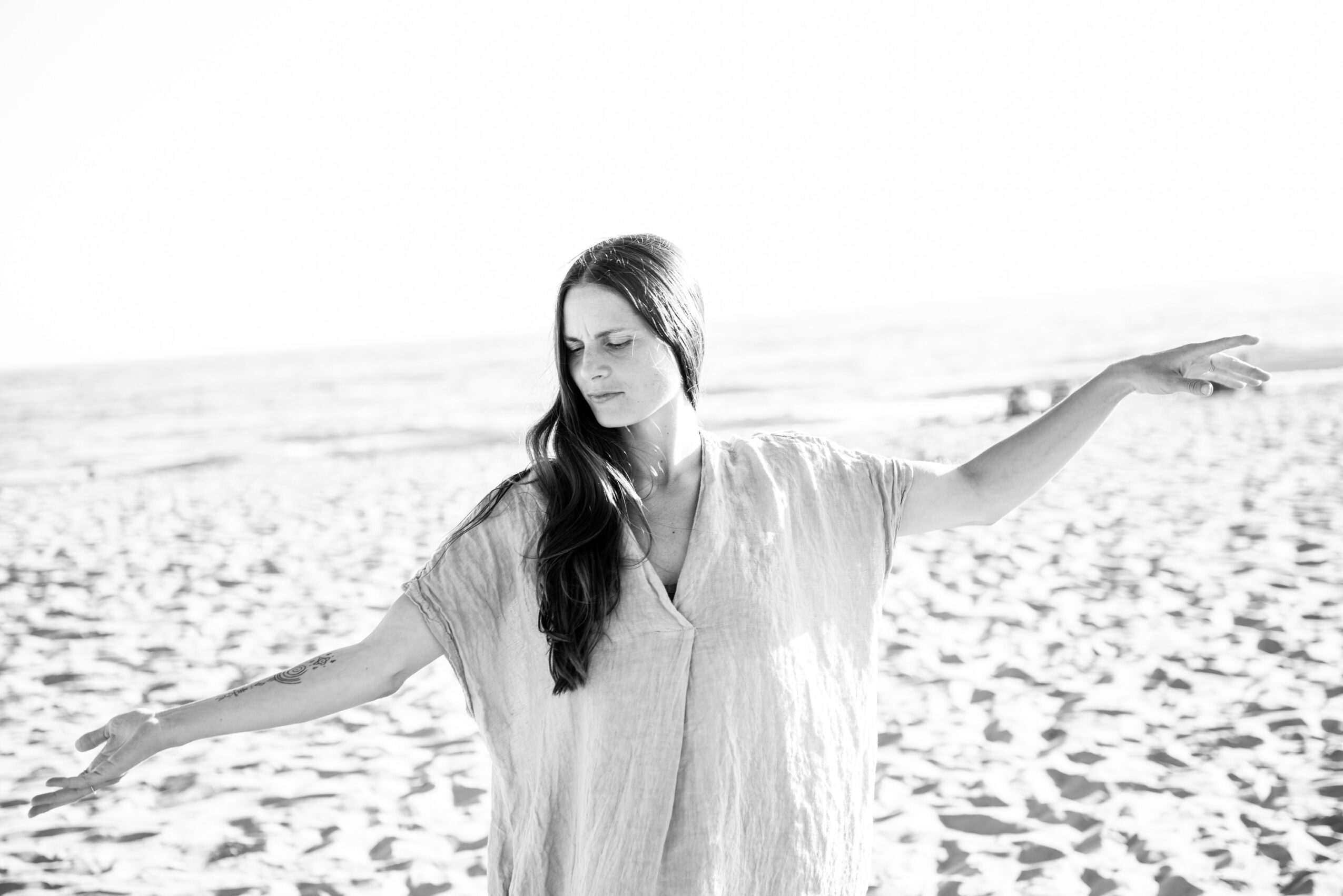 Woman with long hair standing on beach with arms outstretched, facing sideways, in black and white.