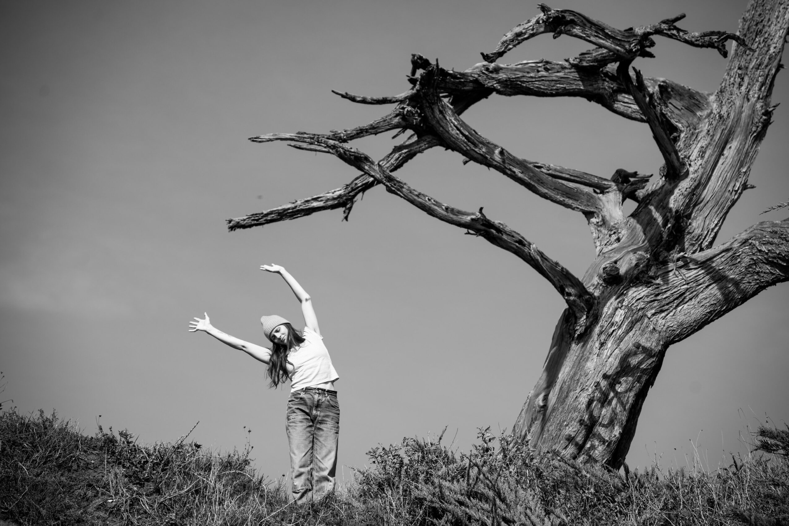 Person standing with arms raised near a large, leafless tree against a cloudy sky, black and white image.