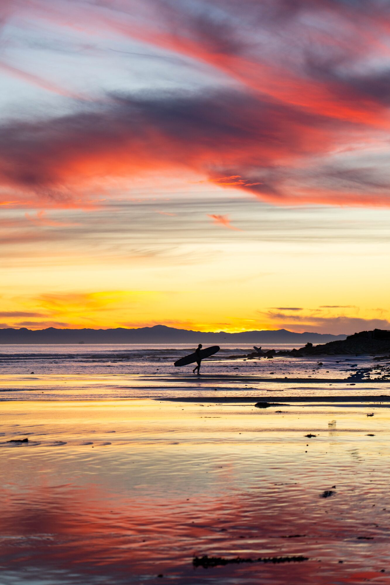 Person carrying a surfboard walking along a beach at sunset with colorful sky and reflections on wet sand.