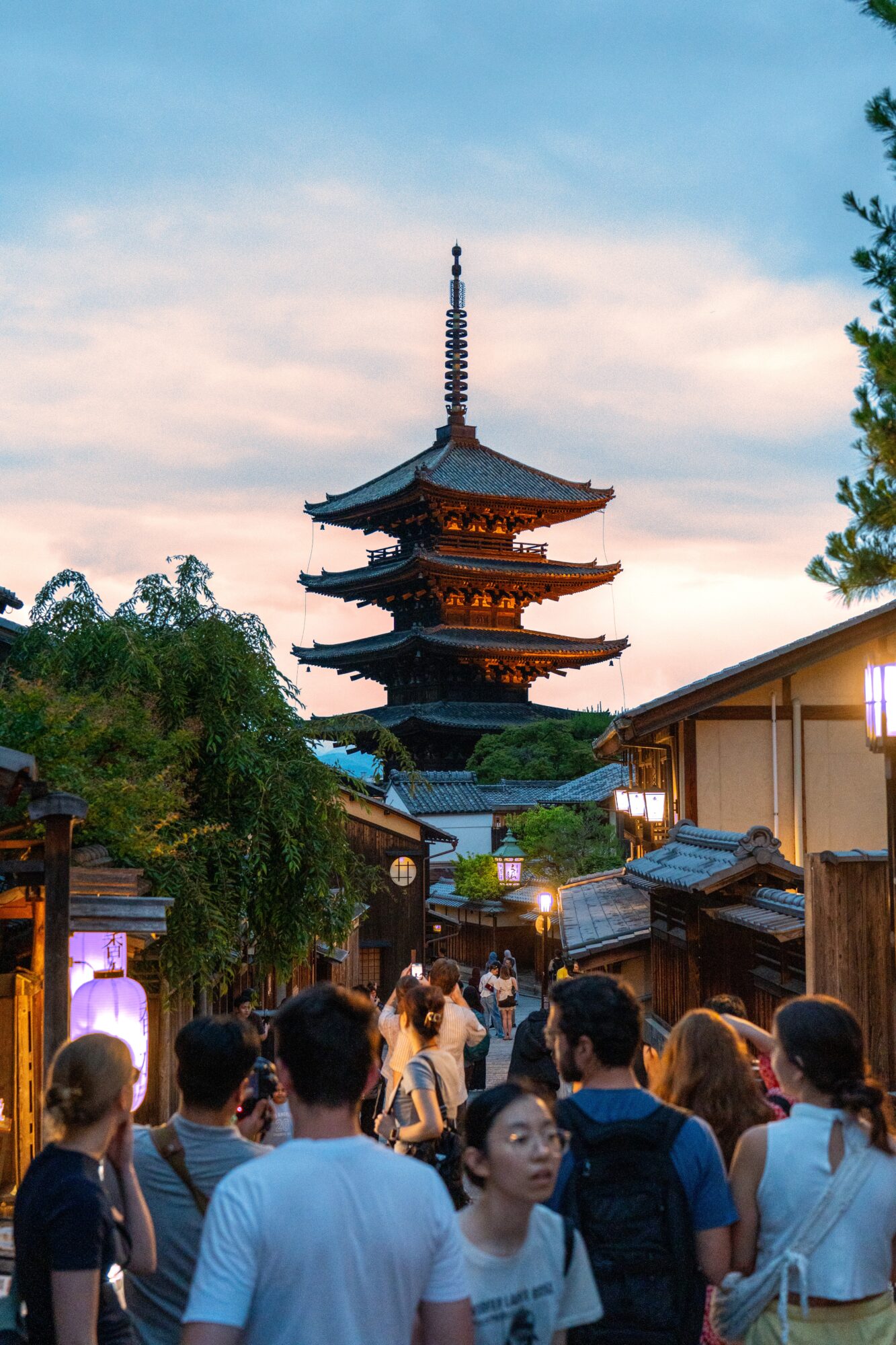 Crowd of people walking in a street with traditional buildings, with a pagoda in the background under a sky with pink and blue clouds.