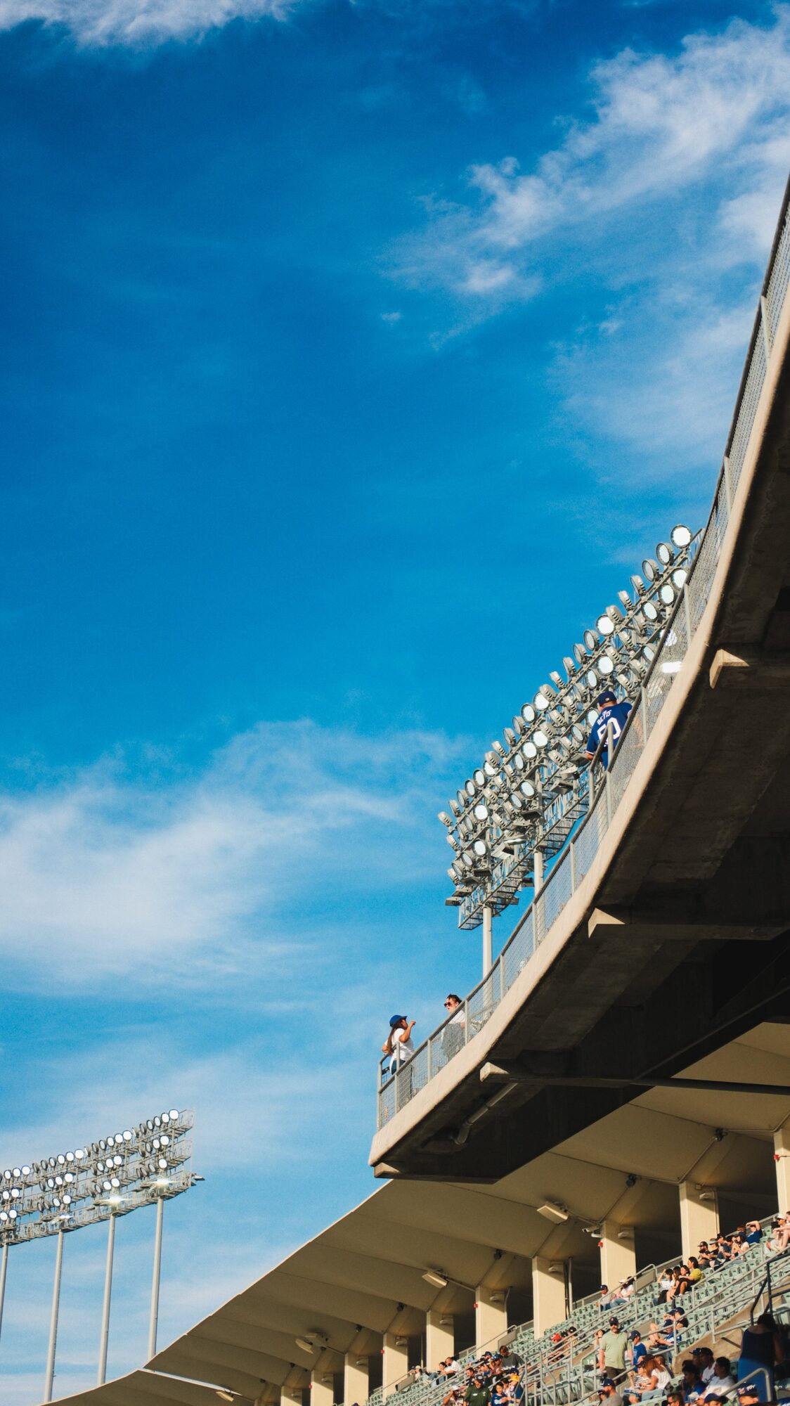 View of a stadium with bright lights and a person standing on the balcony, under a blue sky with clouds.