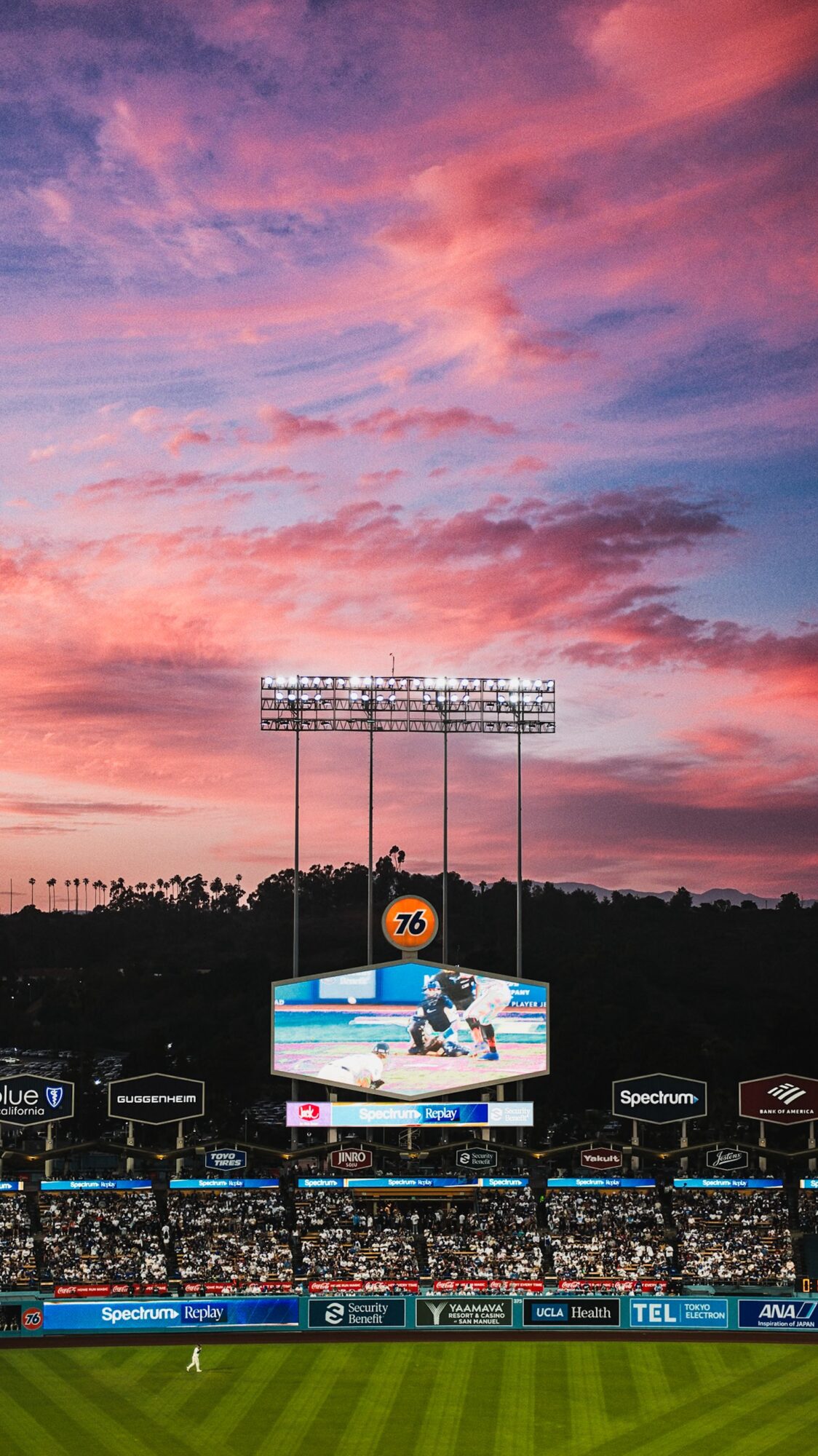Baseball stadium with a large digital scoreboard displaying a player, under a colorful sunset sky.
