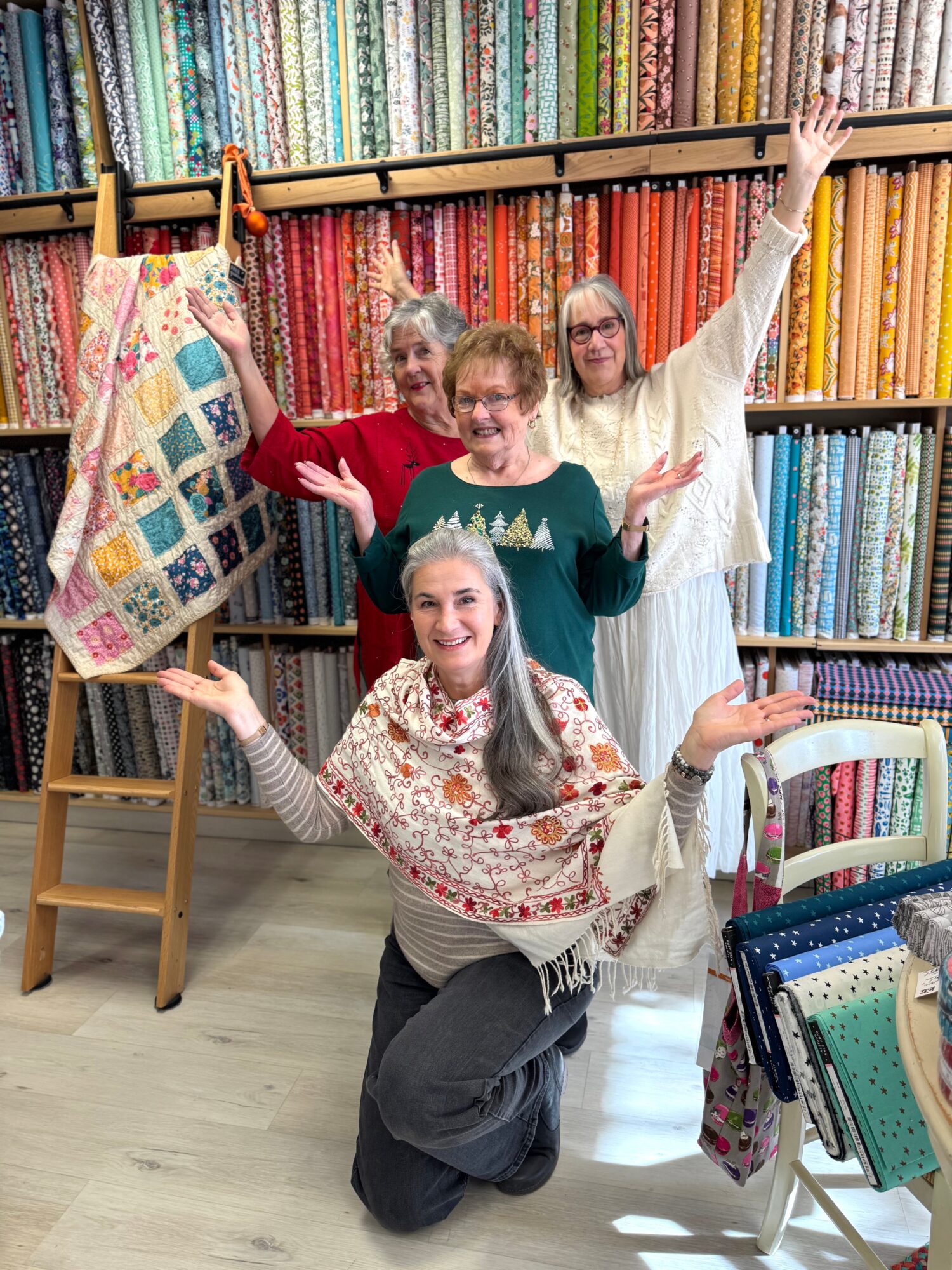 Four women in a fabric store, surrounded by colorful fabric shelves, smiling and posing with arms raised.