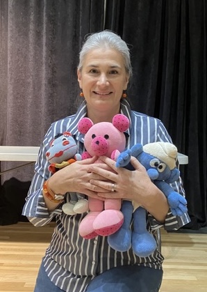 Woman with gray hair holding colorful plush toys, standing in front of dark curtains, smiling.