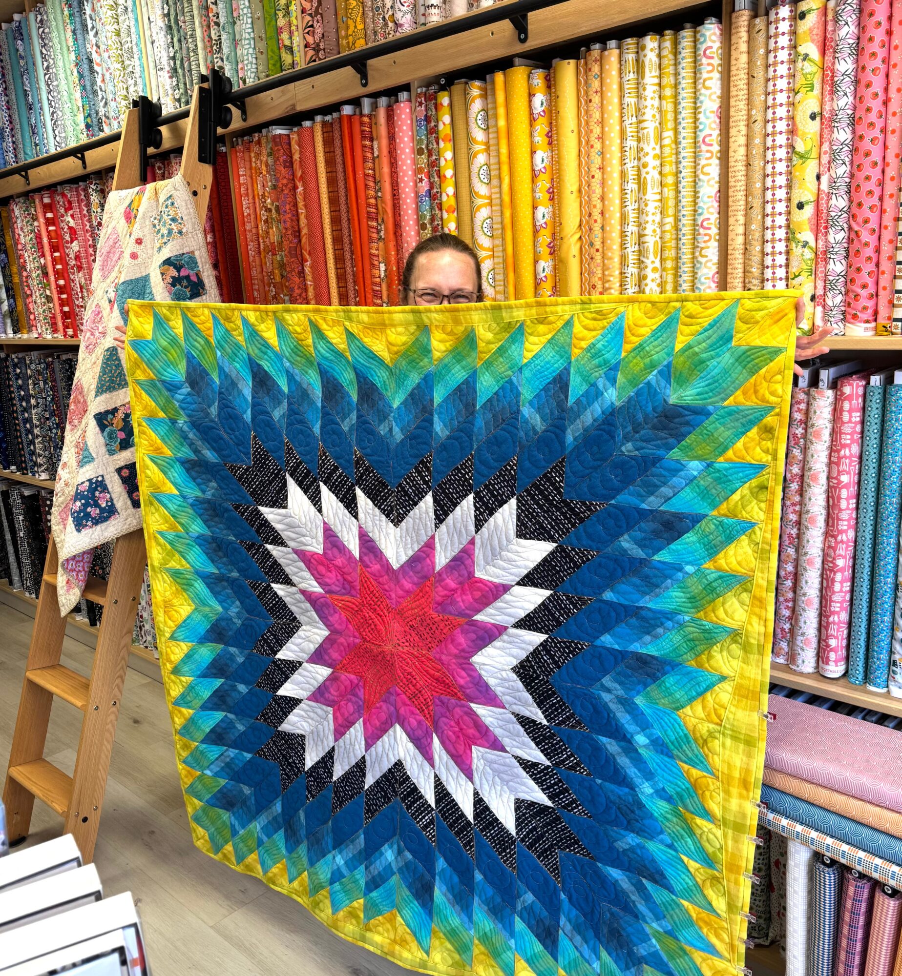 Person holding a colorful quilt with a starburst pattern in a fabric store, shelves of fabric in the background.