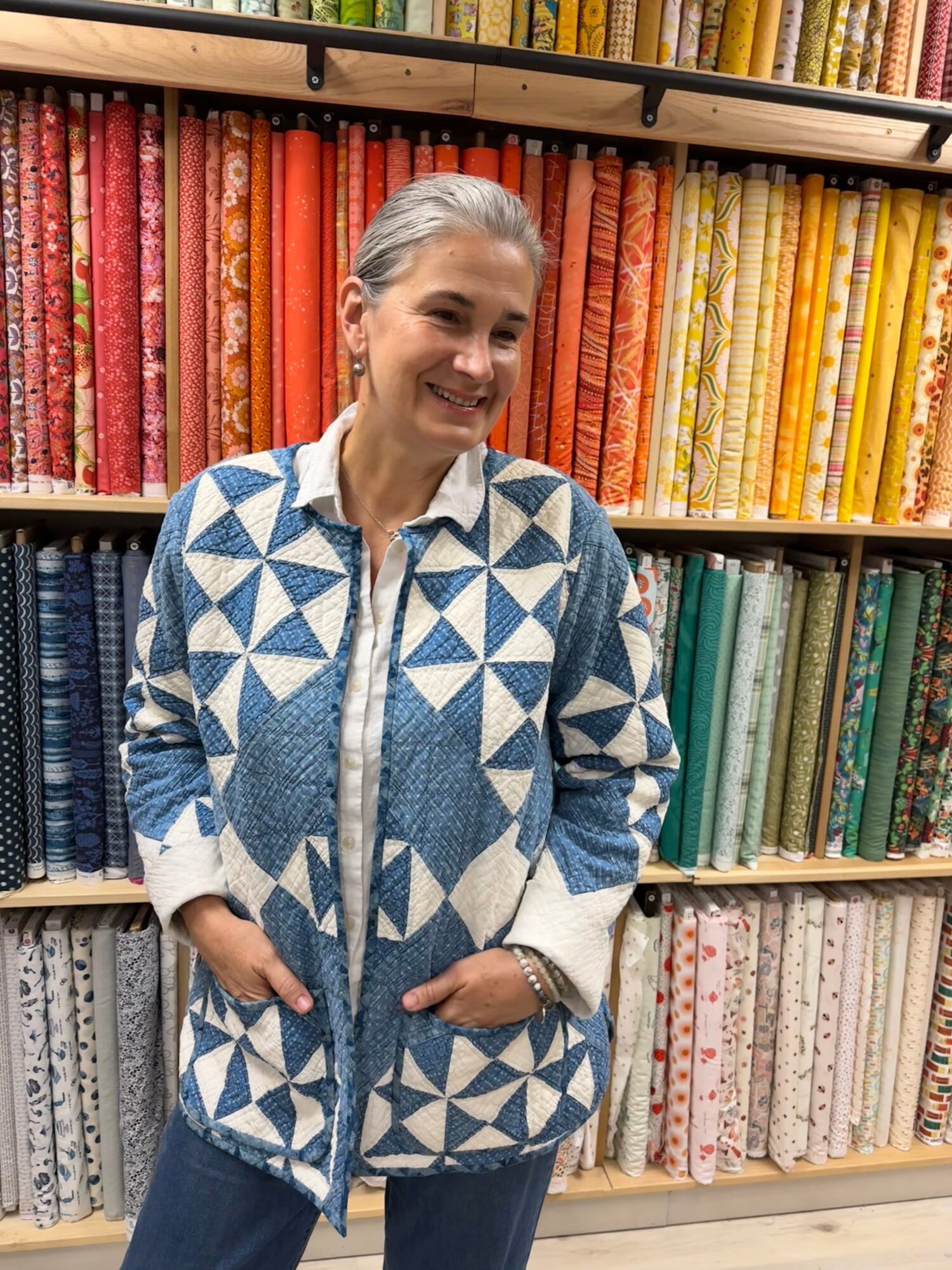 Smiling woman with gray hair in a patterned blue and white jacket stands in front of colorful fabric shelves.