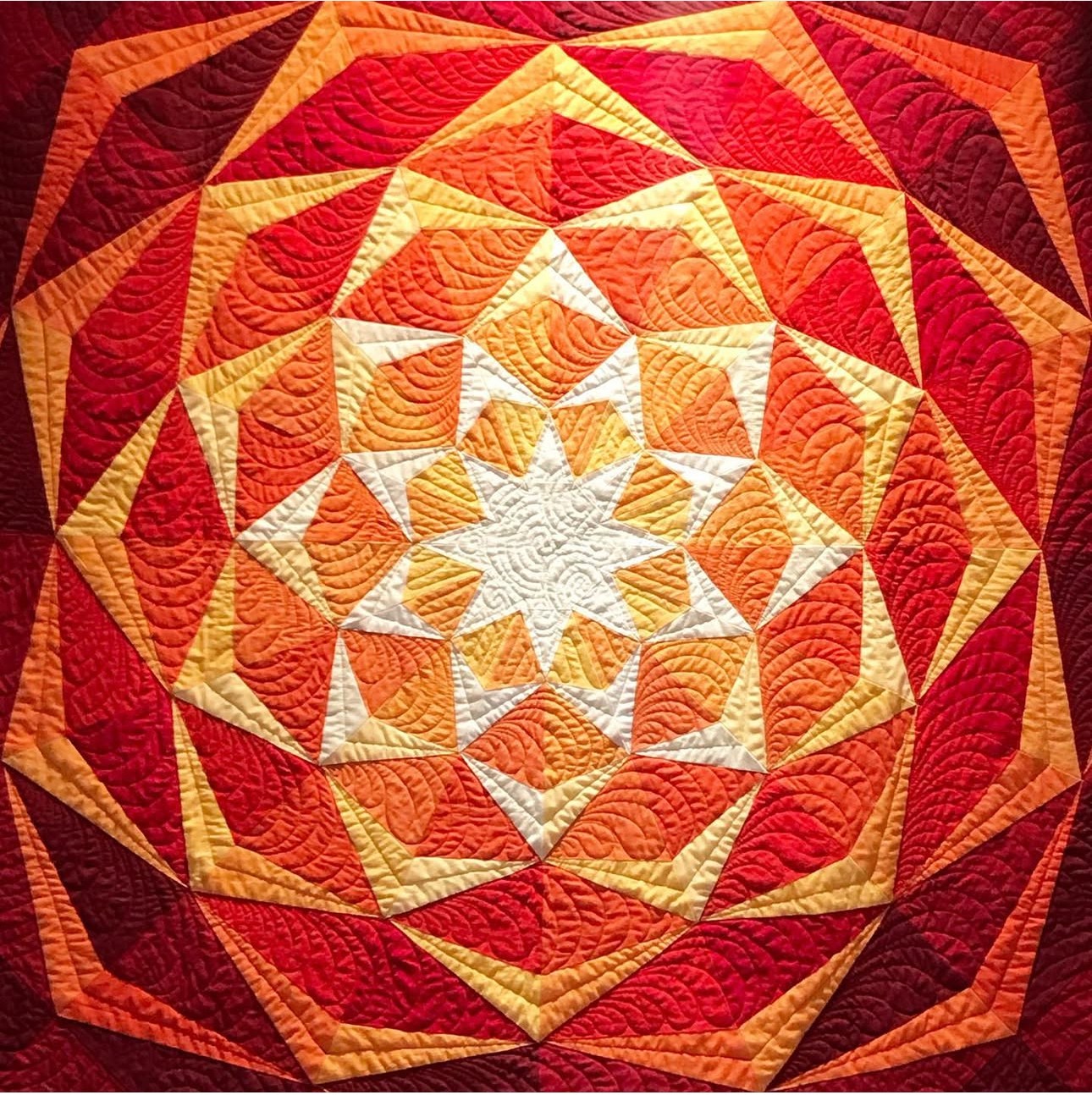 Colorful quilt with geometric star pattern in red, orange, yellow, and cream shades, radiating from the center outward.