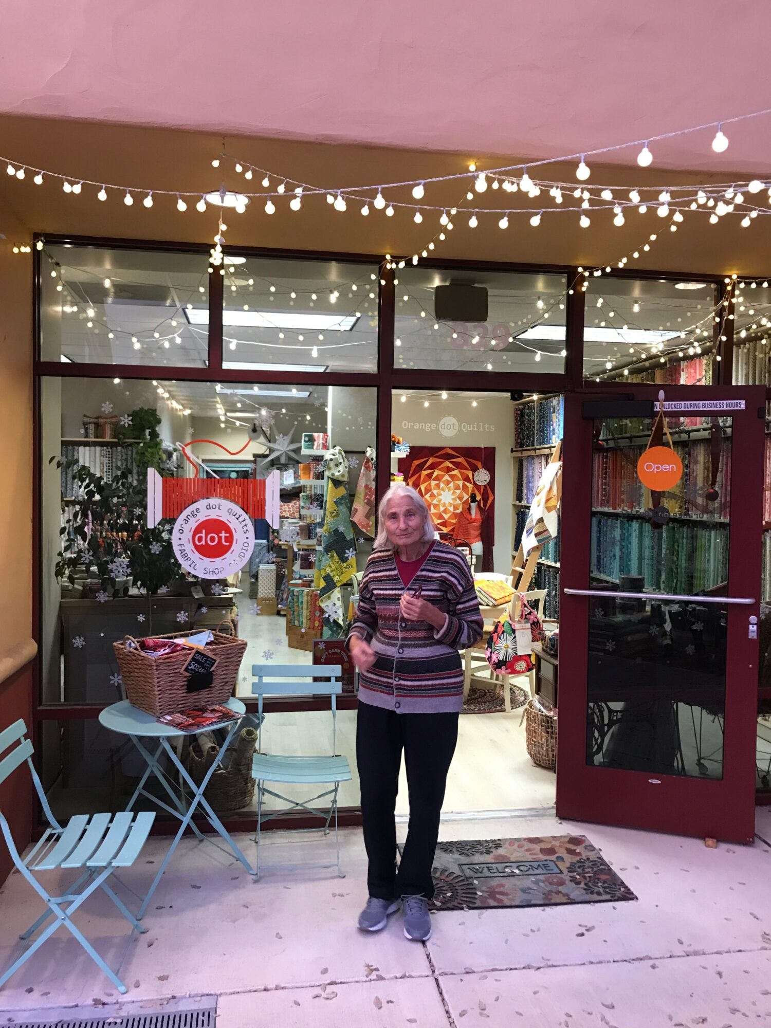 Person standing outside a store with glass windows, string lights, and a red door, holding a bag, with a small chair nearby.
