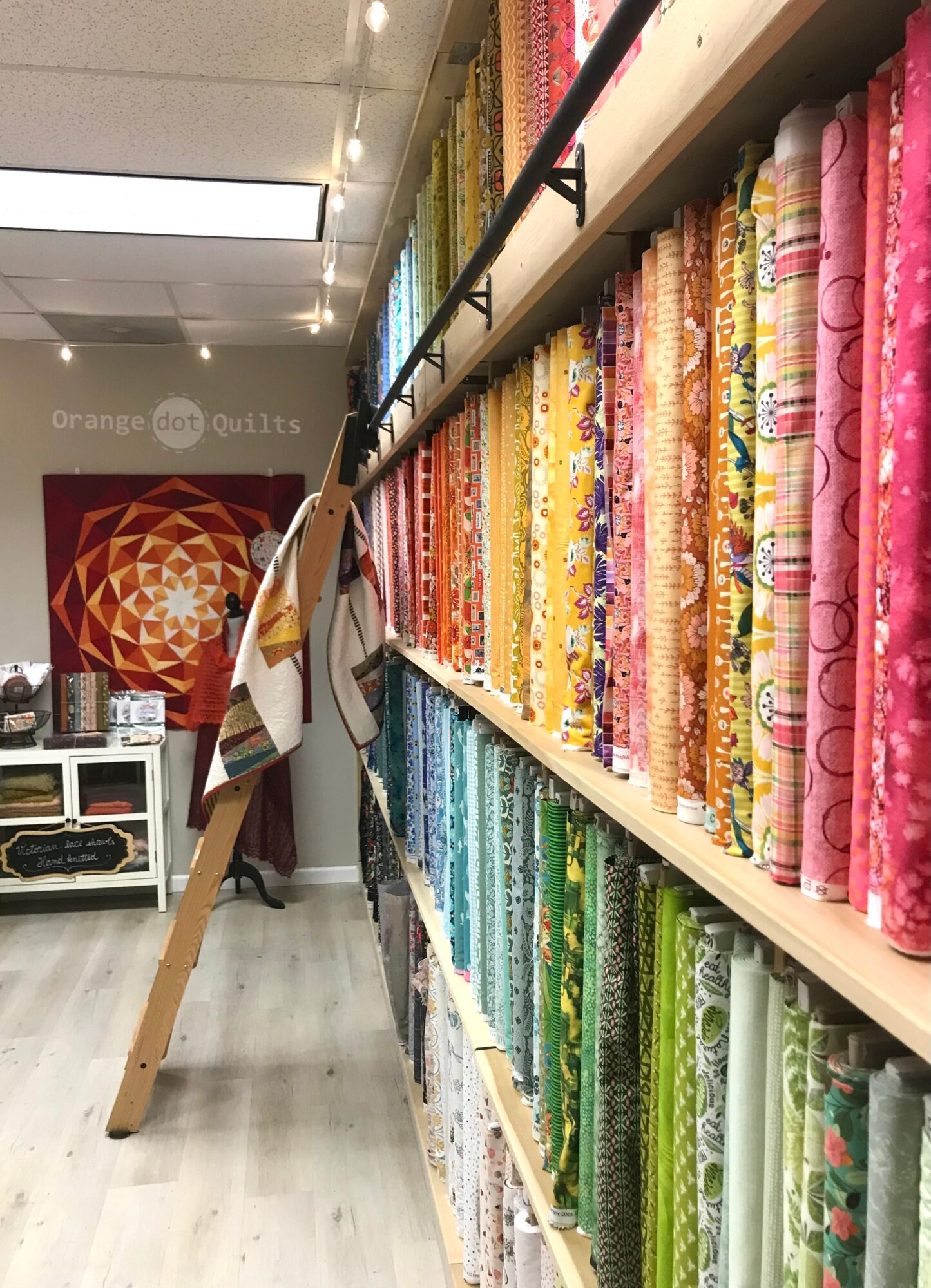Store aisle with shelves of colorful fabric rolls, a wooden ladder leaning, and a person browsing in the background.