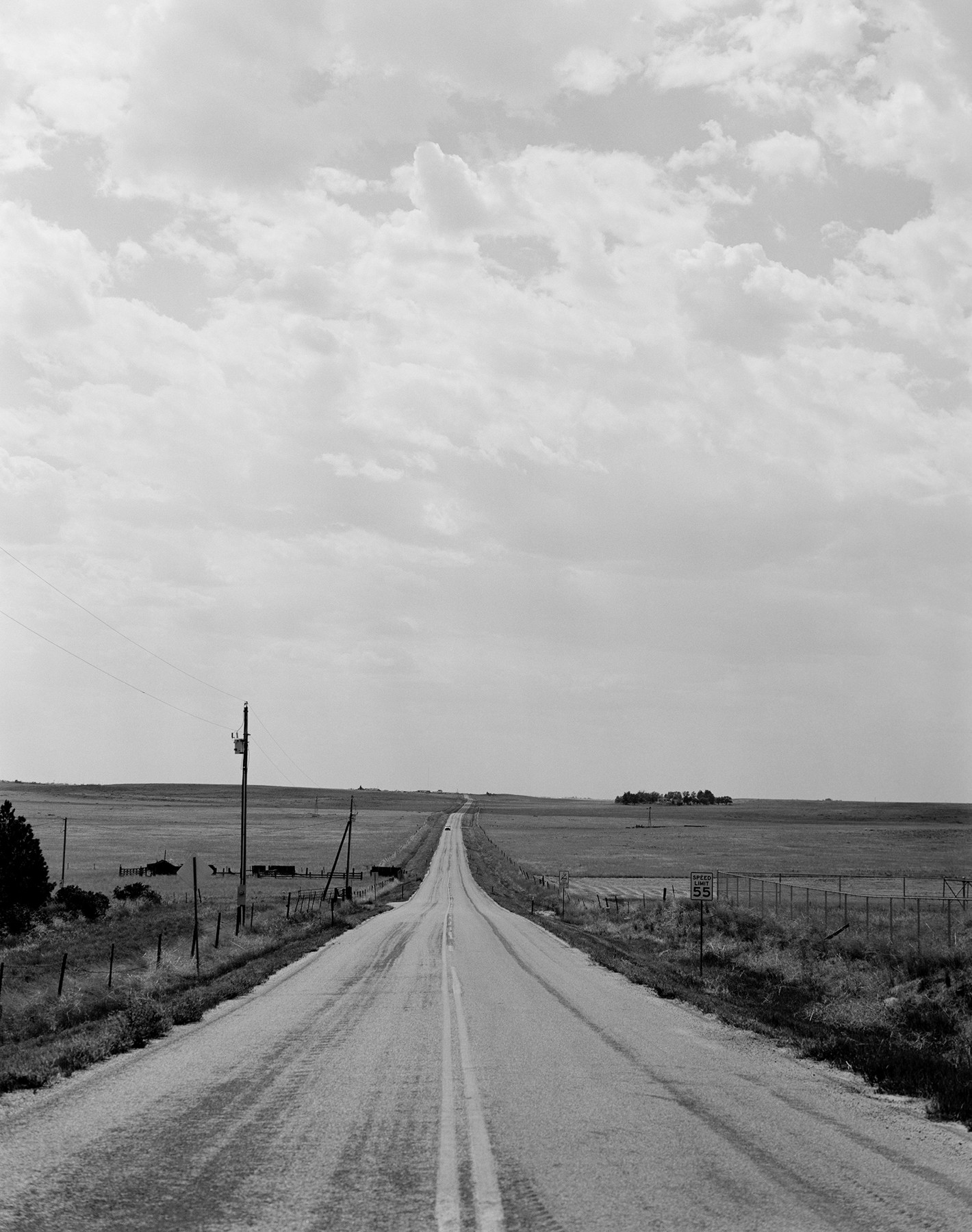 Long straight rural road with double yellow lines, fields on both sides, and a cloudy sky above.