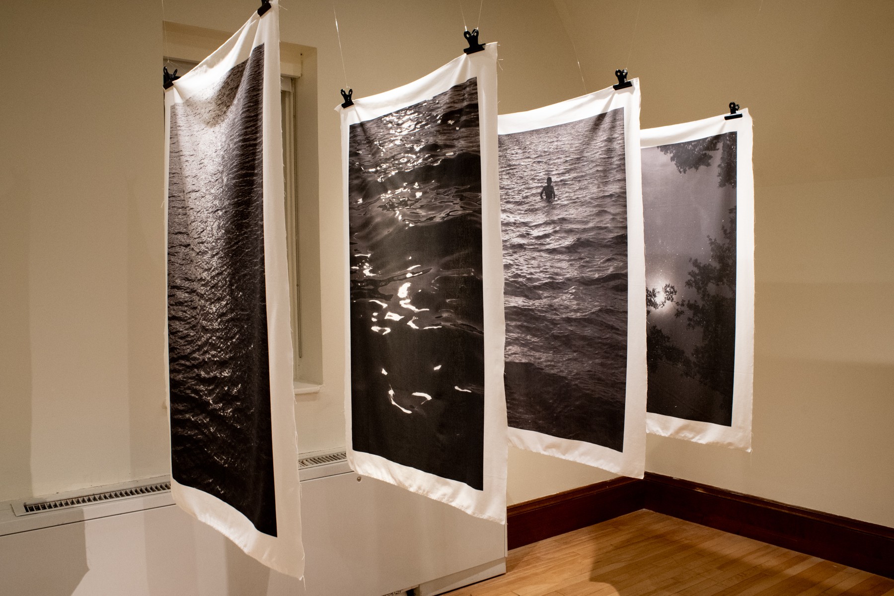Four black-and-white photographs hanging on a wall, depicting water scenes, suspended with clips, with a wooden floor and beige wall.