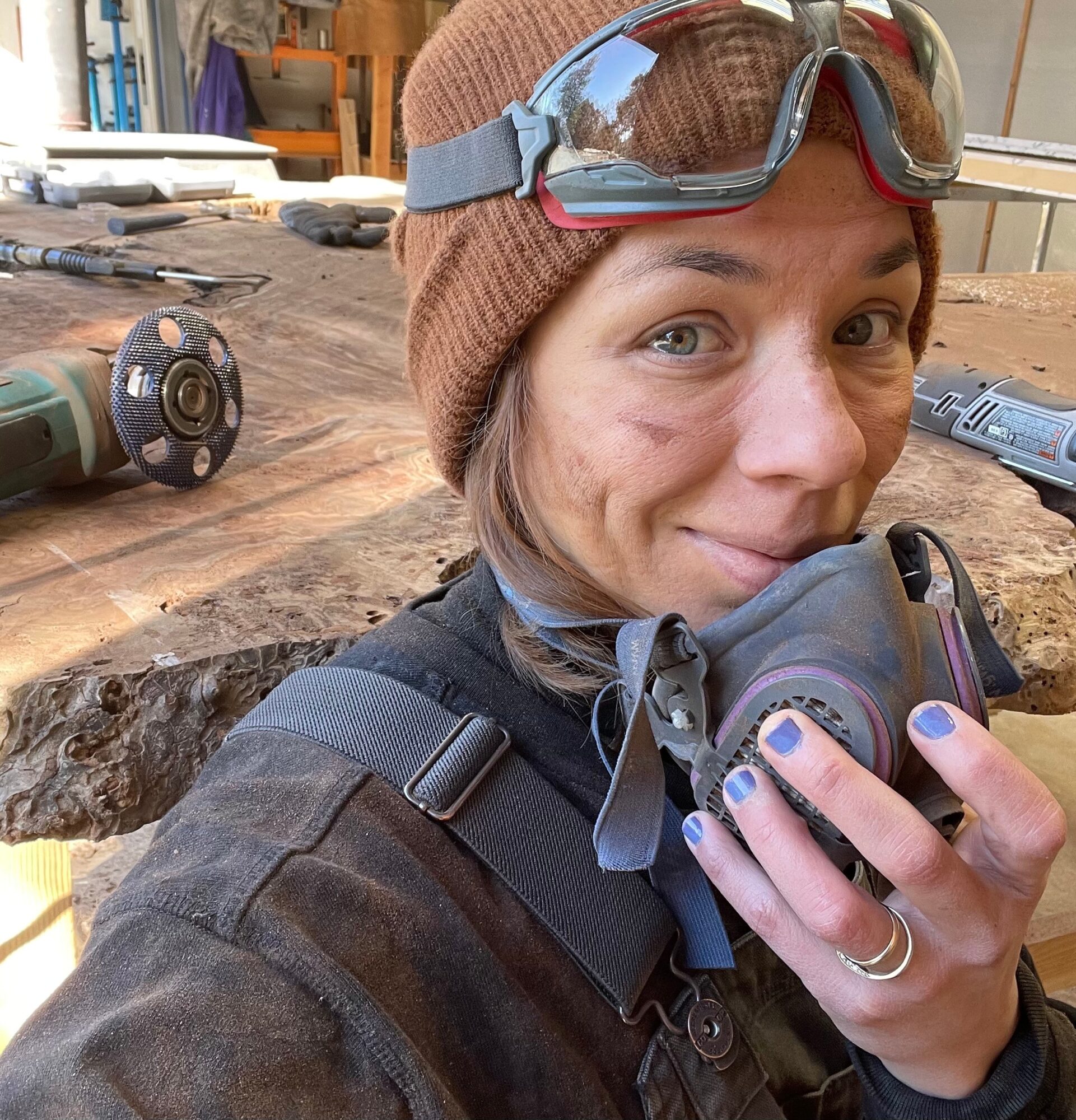 Woman in a workshop holding a respirator, wearing goggles, a beanie, and a black jacket, with woodworking tools in the background.