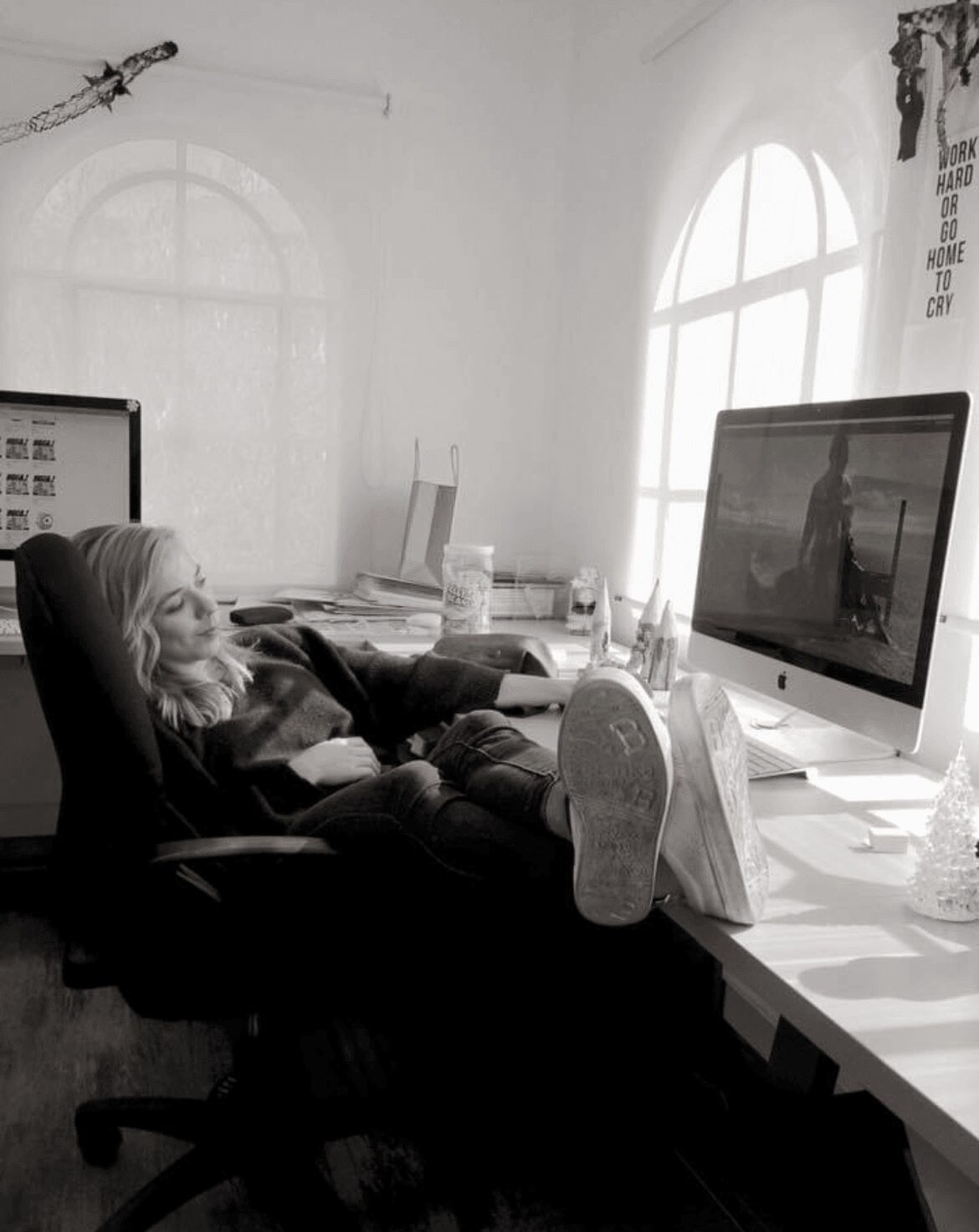 Person resting with feet up on a desk, using a computer in a bright room with large windows.