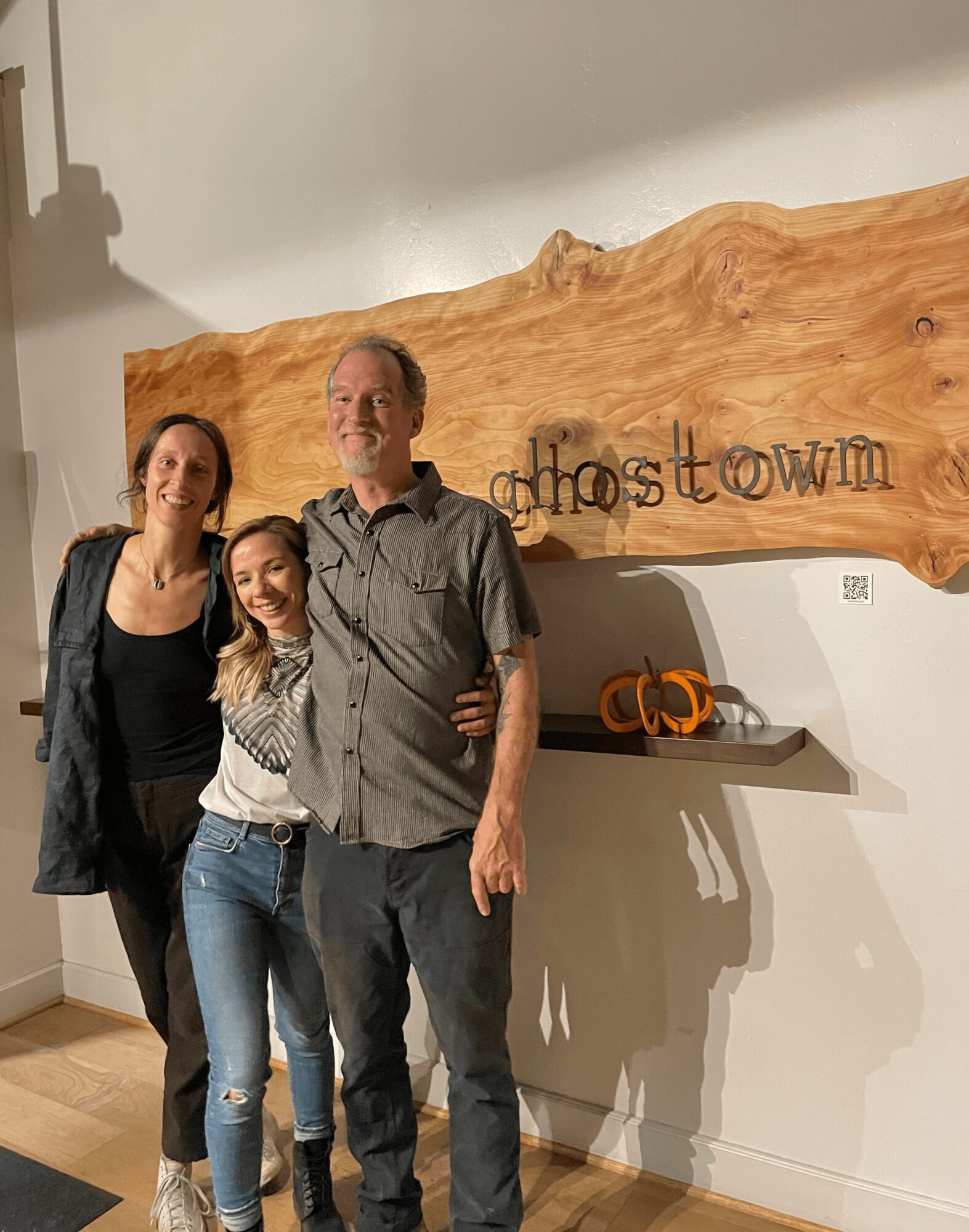 Three people standing together indoors, smiling, with a wooden sign reading 'ghostown' on the wall behind them.