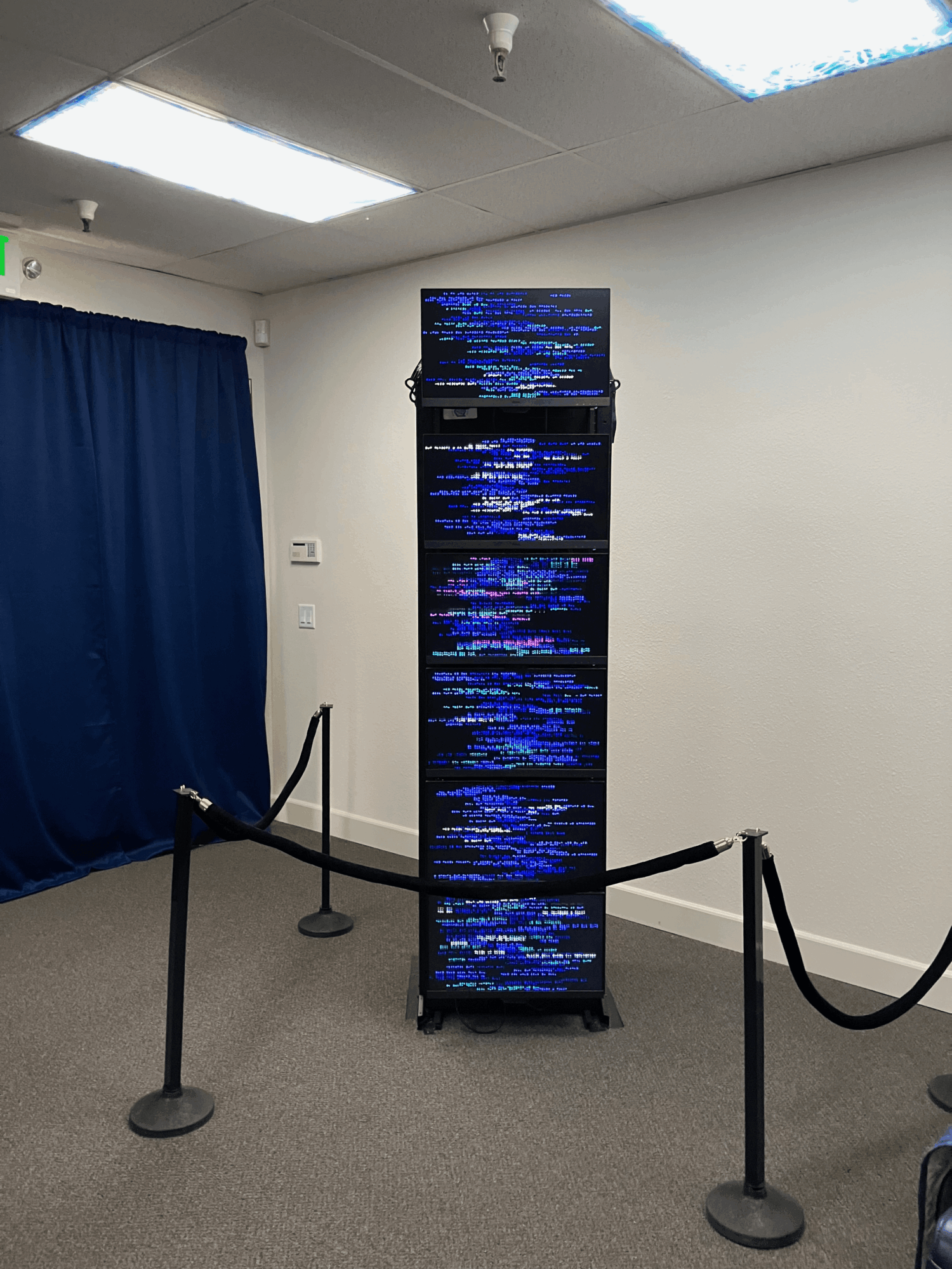 Tall digital display screen with blue and white text, surrounded by black stanchions and ropes, in a room with white walls and ceiling lights.