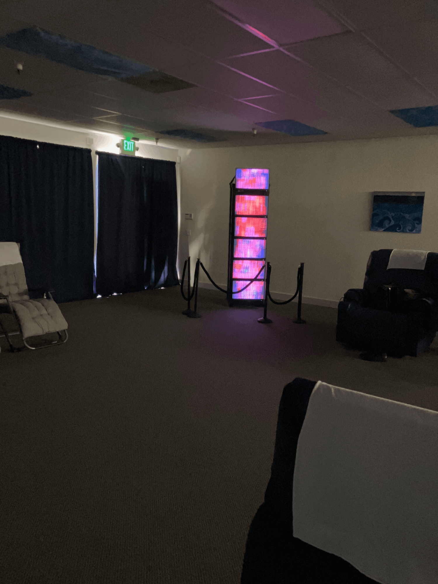 Dimly lit room with a tall, illuminated display, a black sofa, and a curtain-covered window or door, with a chair in the foreground.