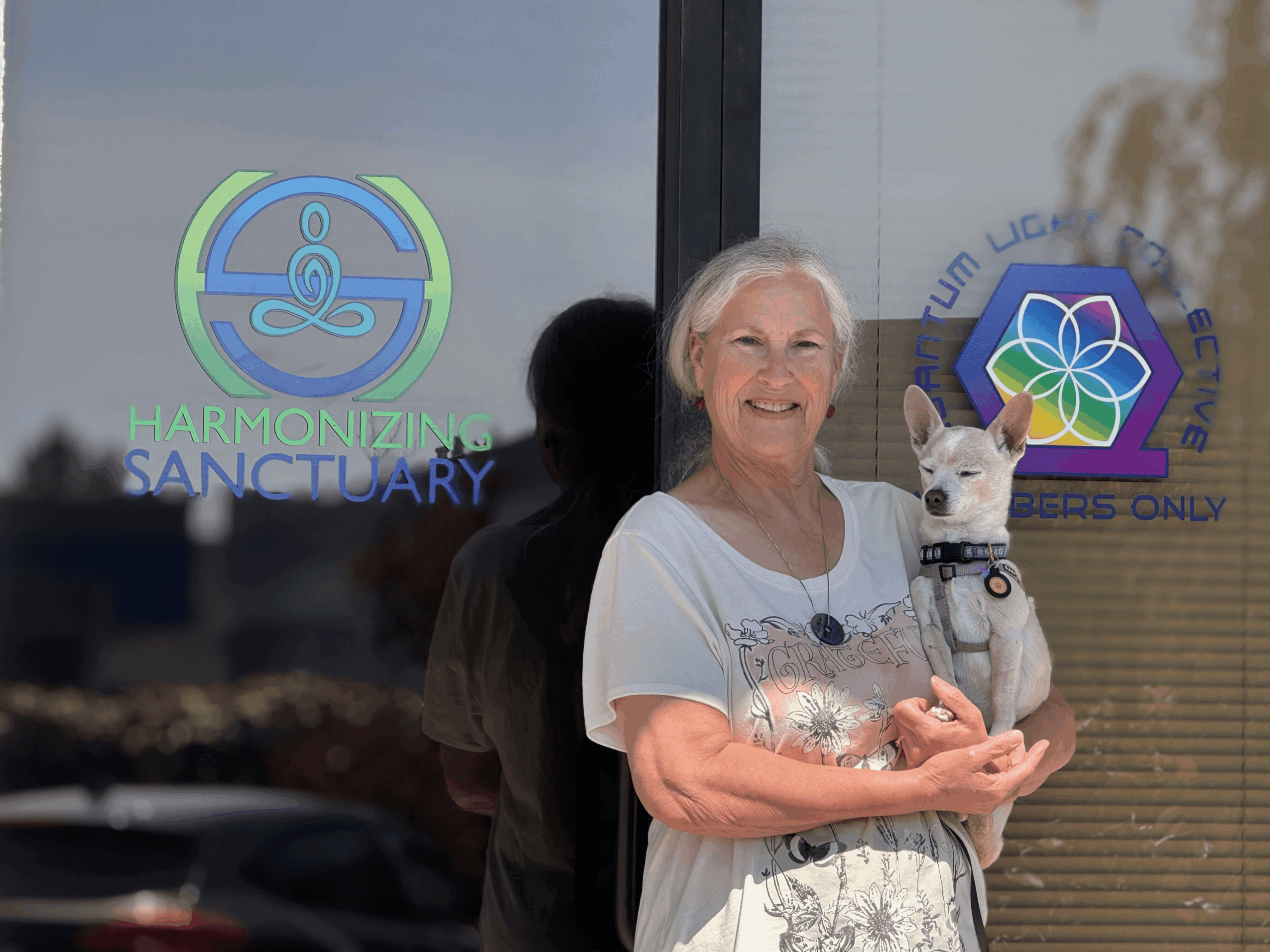 Smiling woman holding a small dog outside a building with signs for Harmonizing Sanctuary and Therapy Dogs Only.