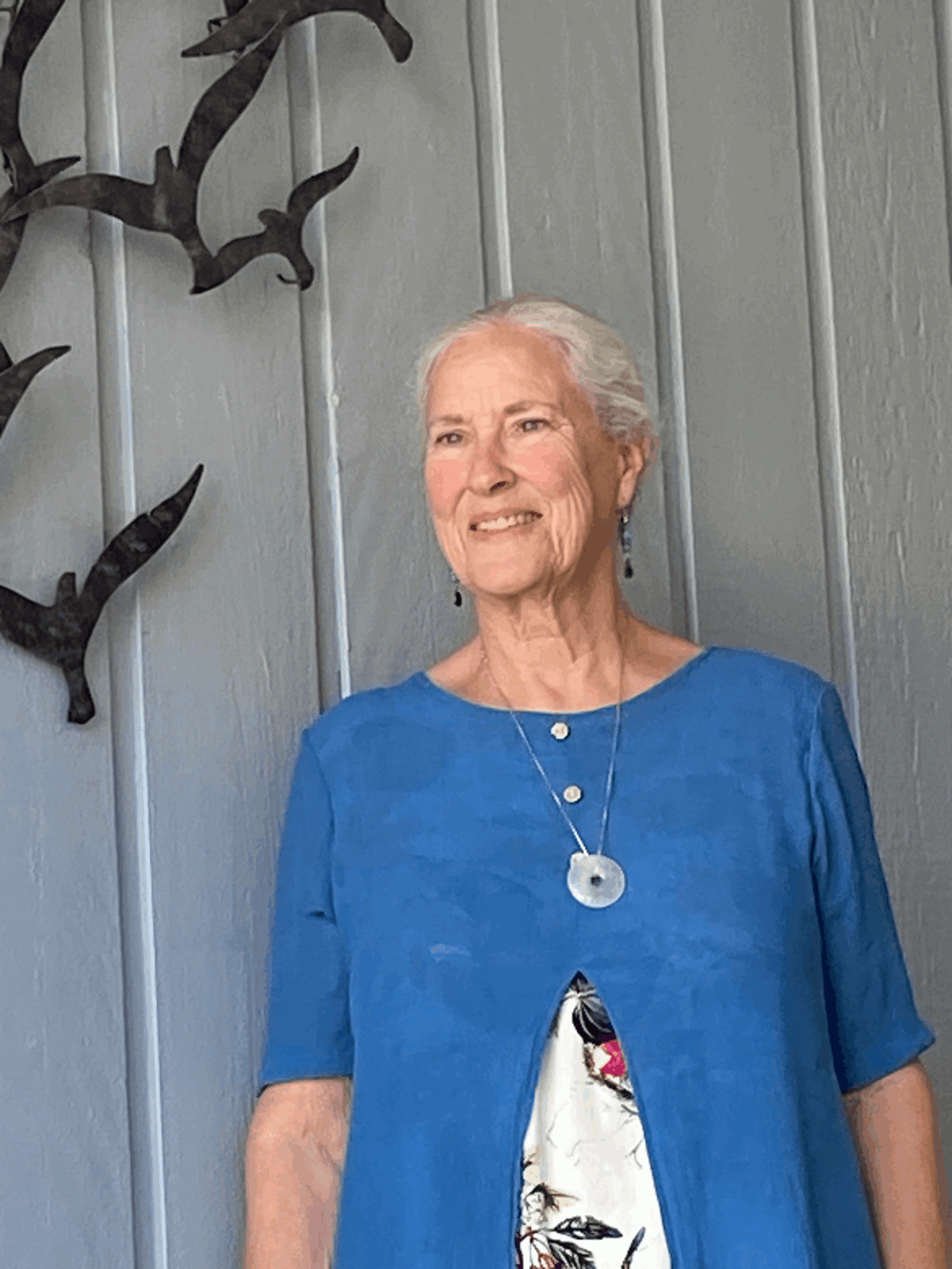 Elderly woman with white hair wearing a blue dress and necklace, standing outdoors near a gray wall.