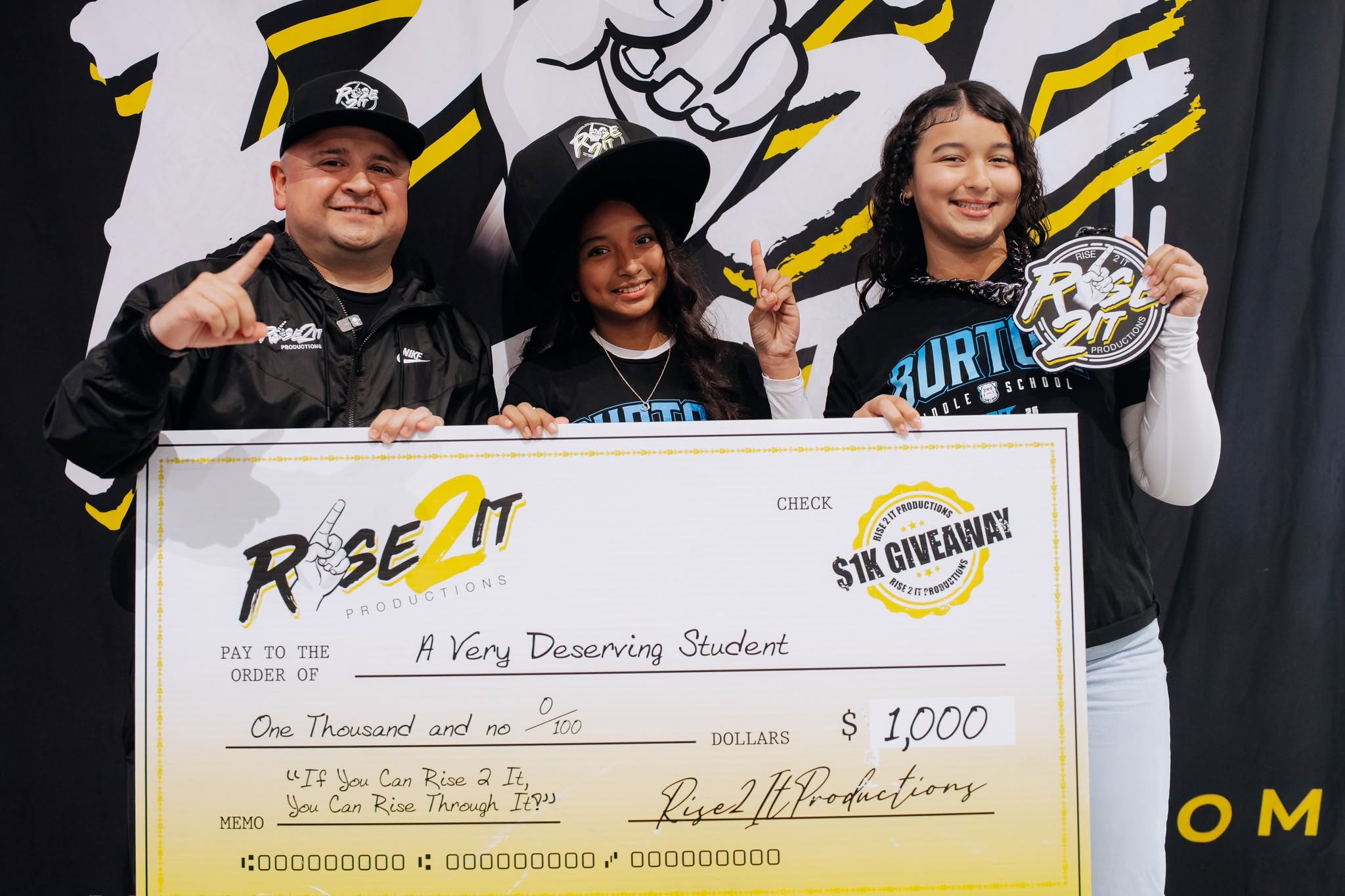 Three students holding a large check, smiling, in front of a black and yellow backdrop, celebrating a scholarship award.