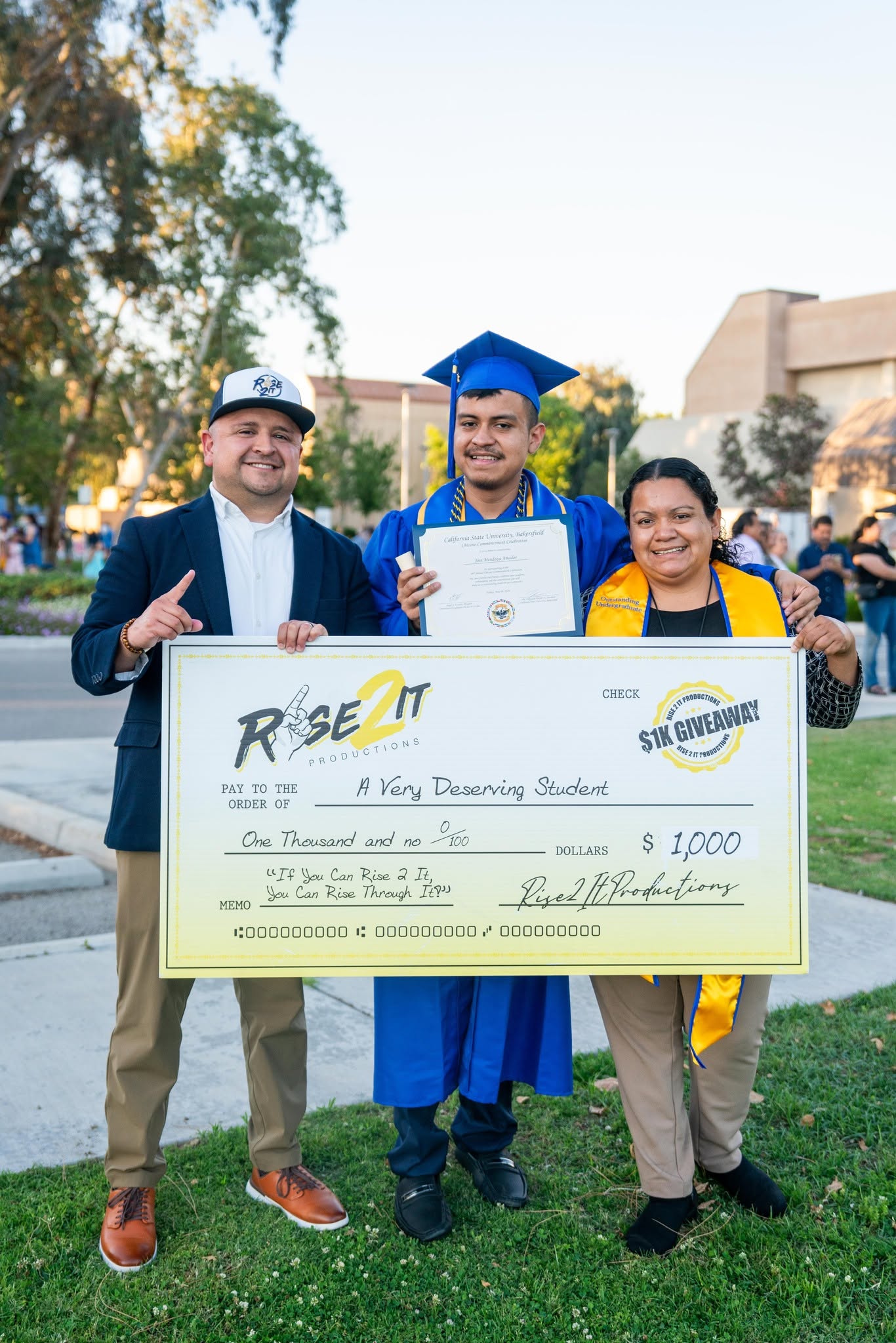 Three people, one in a graduation cap and gown, holding a large check outdoors on grass, smiling, with trees and buildings in background.