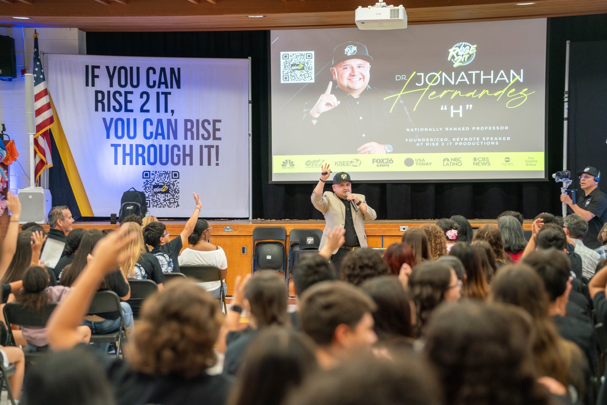 Speaker on stage in front of audience, presentation slide with motivational quote, audience raising hands, flags on side of stage.