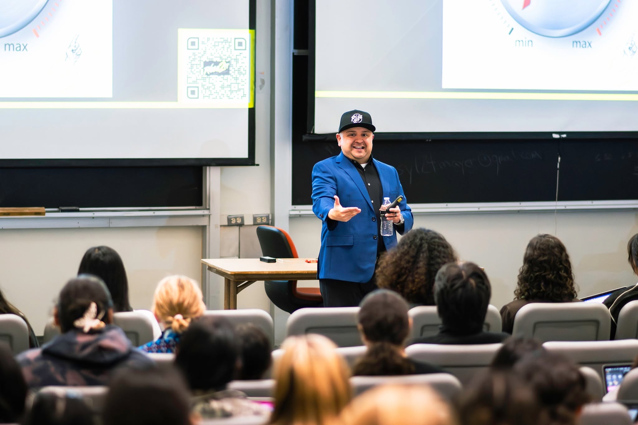 Person in blue jacket and cap speaking to an audience in a classroom or conference room, with presentation screens behind.