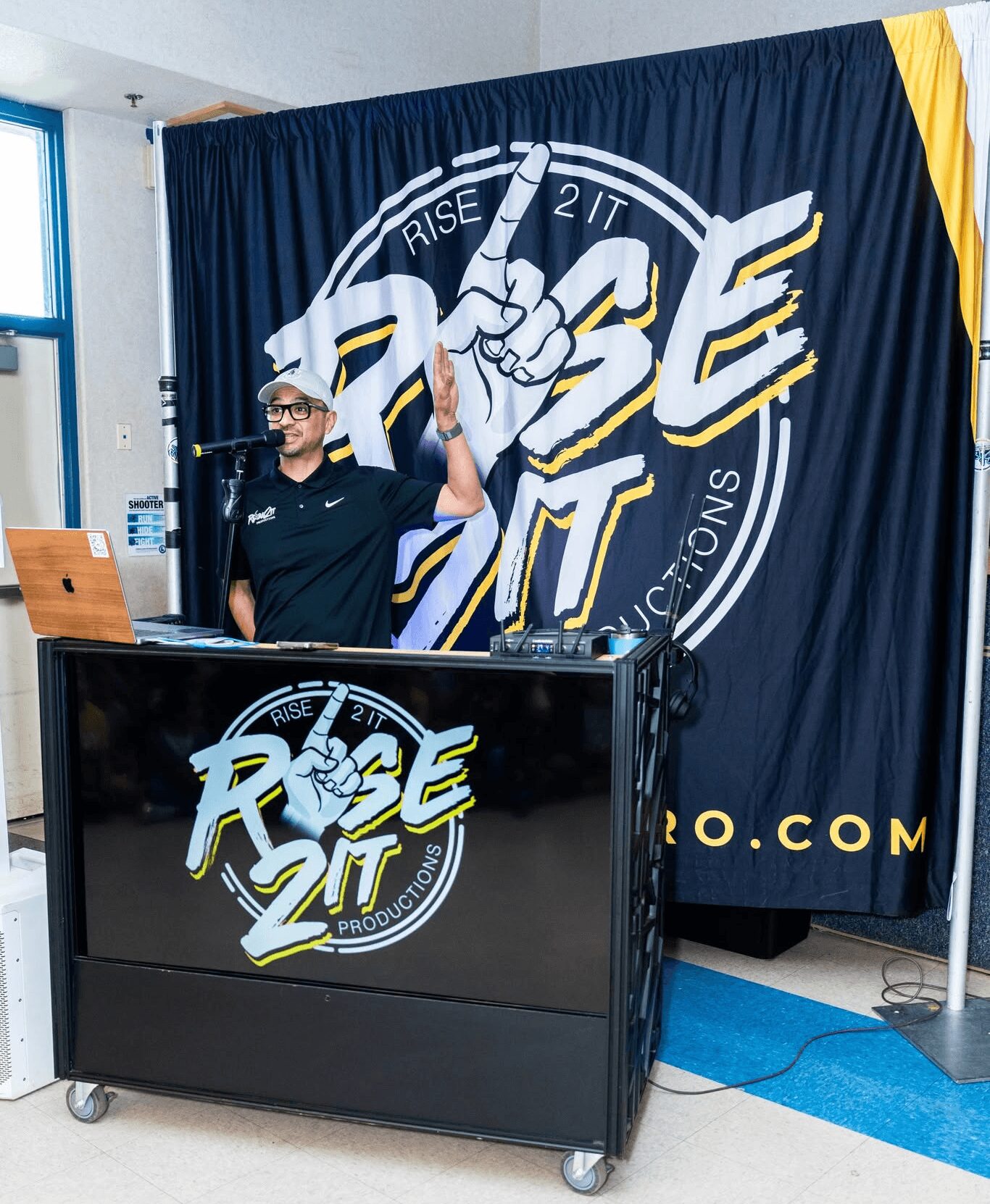 Person standing behind a DJ booth with a Rise Up logo, holding a hand sign, in front of a large Rise Up banner.