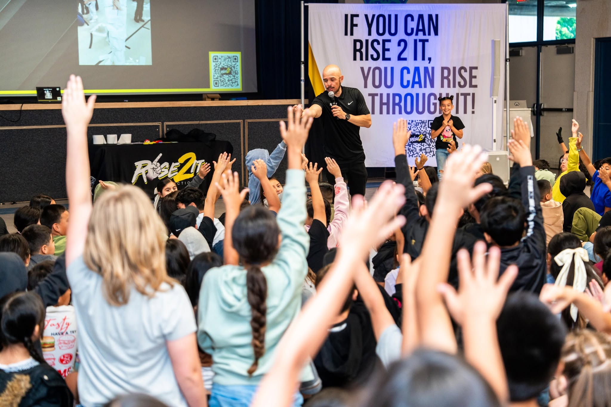 Crowd of children raising hands in front of a stage with speakers and a large screen displaying motivational text.